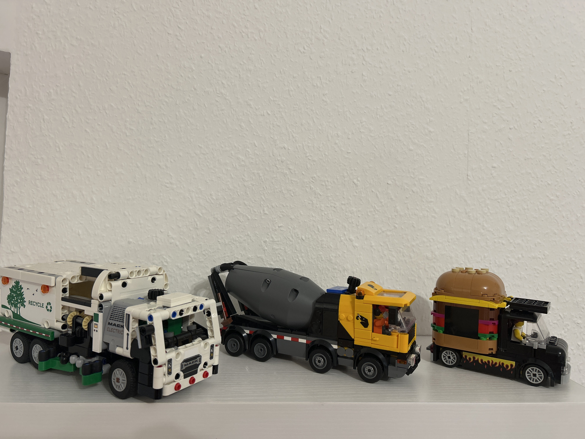 The image shows three Lego trucks on a white shelf in front of a white textured wall. The leftmost truck is a white garbage truck with green accents and the word "RECYCLE" printed on its side. The middle truck is a yellow and black cement mixer. The rightmost truck is a black food truck with a burger on top. The wall is plain and undecorated. The lighting appears to be from a single source, casting soft shadows. There are no people visible in the image. The scene is static and appears to be a display.