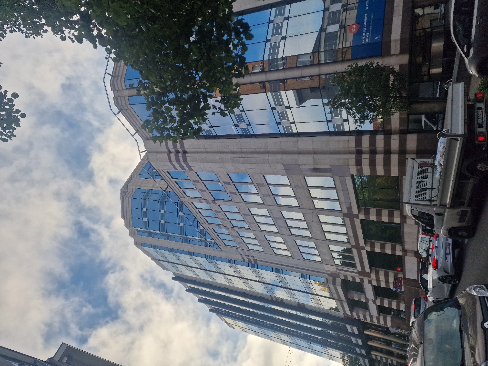 This is a ground-level, low-angle shot of a modern, multi-story office building with a glass facade reflecting the sky and surrounding buildings. The building has beige stone accents with dark horizontal stripes. In the foreground, there is a traffic jam on a street, with several cars visible, including a white convertible and a light-colored truck. Trees are present on the right side of the image, partially obscuring the building. The sky is partly cloudy with patches of blue visible. A blue banner with white text is attached to the building on the right side, displaying "LA RÉGIE DES BÂTIMENTS ENGAGE!" and "DE REGULIERE. A VOUS VOTRE COLLEGE !". This scene is located in Saint-Gilles - Sint-Gillis, Belgium.