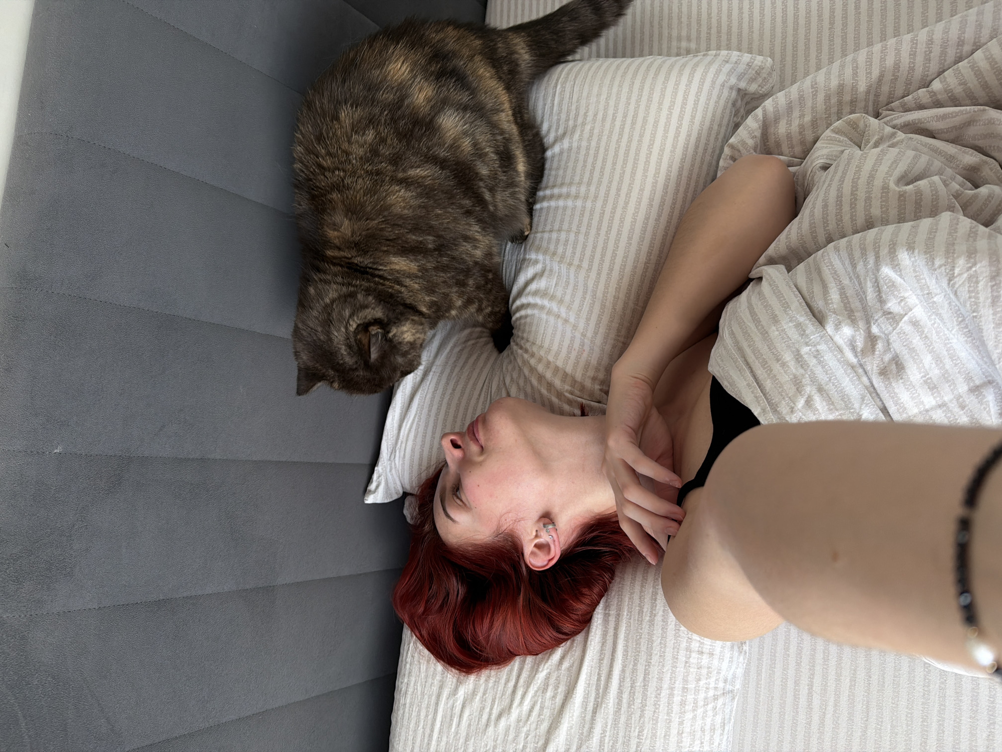 A high-angle shot depicts a woman with red hair lying on a bed with striped bedding, with a tabby cat standing near her head on the same pillow. The woman is fair-skinned, and partially covered by a duvet. She wears a black top and a beaded bracelet on her extended arm. The bed is positioned against a gray, upholstered headboard. The location is unknown.
