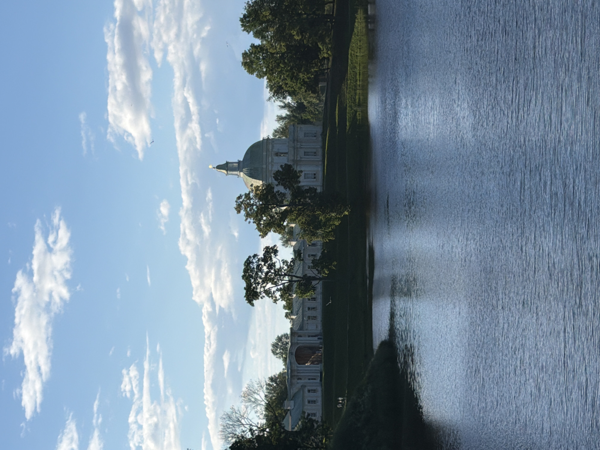 A waterfront view of a neoclassical white building with a green dome and a golden spire, set against a blue sky with scattered white clouds. The building is surrounded by lush green trees and is situated on a gentle slope leading down to a calm body of water. Several small figures are visible on the grounds in front of the building. The water in the foreground reflects the sky and the surrounding greenery.
