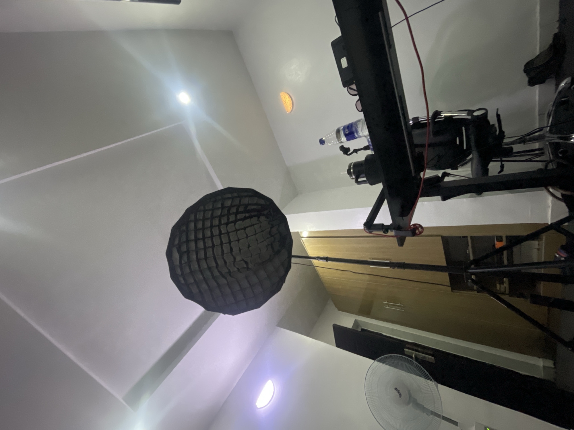 The image depicts the interior of a room, likely in Iddo Tudunwada, Nigeria, given the prompt. The perspective is low, looking upwards. The scene is illuminated by overhead lighting, some of which appears to be from recessed fixtures. A large, dark grid-like modifier, likely for a studio light, is prominently featured. To the right, a black studio boom arm supports a camera and other equipment. A water bottle rests on the arm. In the background, there's a wooden cabinet, a door, and an electric fan. The walls are painted a light color, possibly white or off-white. The room appears to be a small studio or workspace.