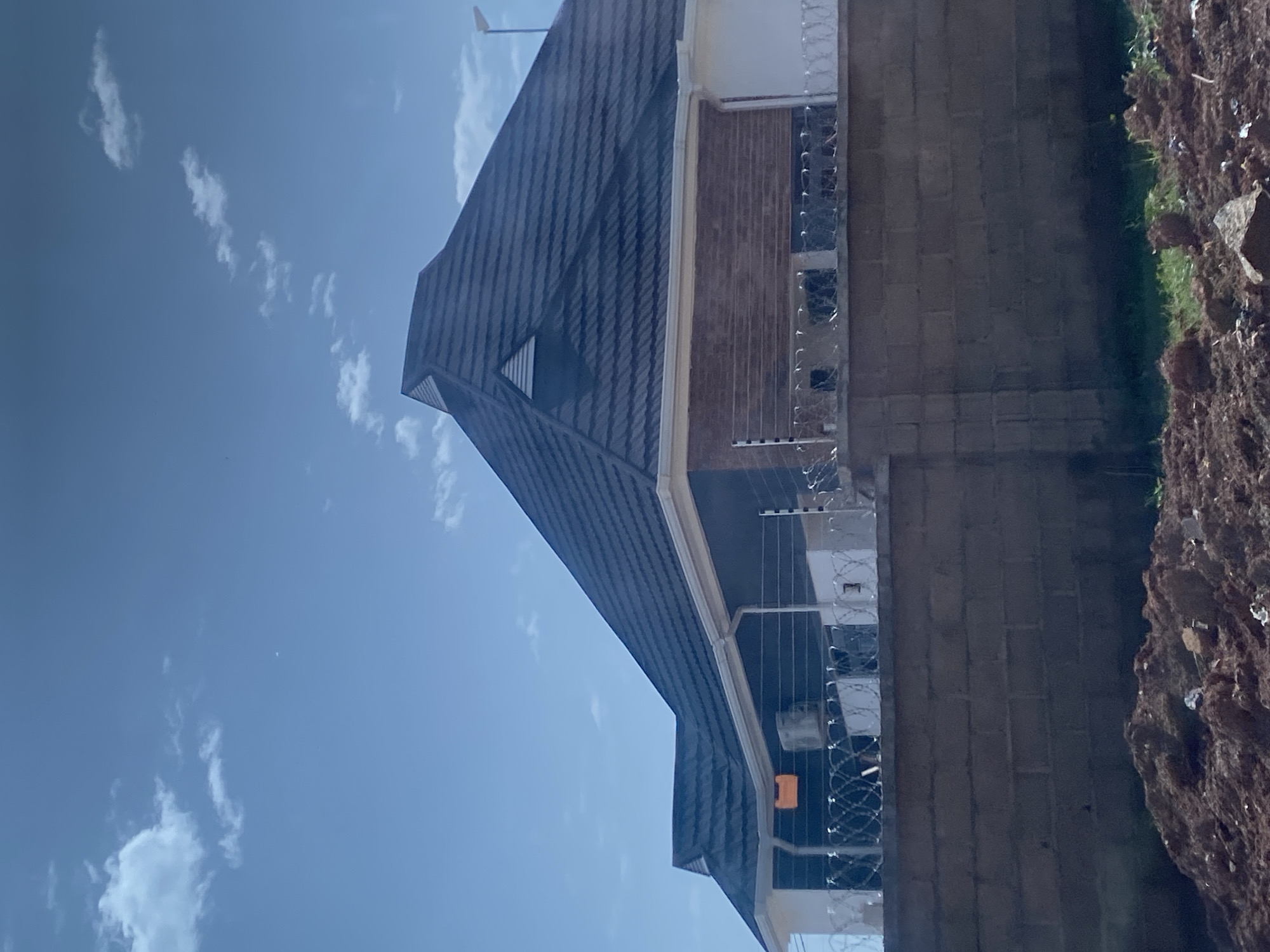 The image shows a modern house with a dark, pitched roof in Bar Jirgi Summa, Nigeria. The house is partially visible, with its upper floors featuring dark siding and a section with a brick-like texture. A white trim runs along the eaves. The property is surrounded by a brick wall topped with barbed wire, suggesting security measures. An air conditioning unit is visible on the side of the house. The foreground of the image is dominated by a pile of reddish-brown dirt and some sparse green grass. The sky above is a clear blue with a few scattered white clouds. The overall scene depicts a residential structure in a semi-developed area.