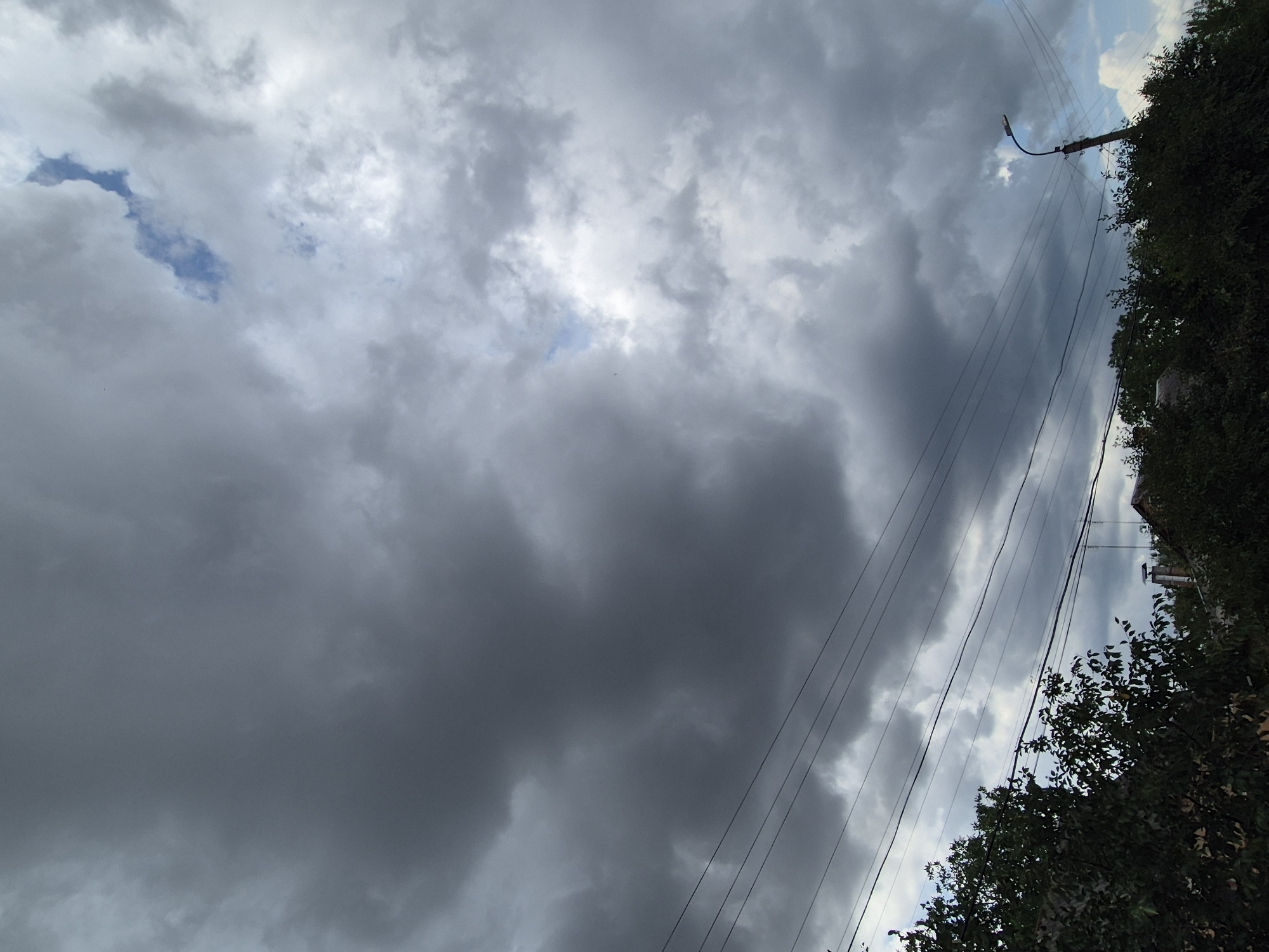 The image depicts an overcast sky with heavy, dark clouds dominating the upper portion. Some patches of lighter sky and a sliver of blue are visible towards the top right. Several parallel power lines stretch across the mid-section of the frame, running from left to right. Below the power lines, the tops of dark green trees and hints of rooftops are visible, suggesting a residential or semi-rural area. A streetlight on a pole is situated on the right side, also partially obscured by the trees. The scene appears to be captured in Sozonivka, Ukraine, under what seems to be a prelude to or aftermath of rain, given the dense cloud cover. No people or specific actions are discernible.