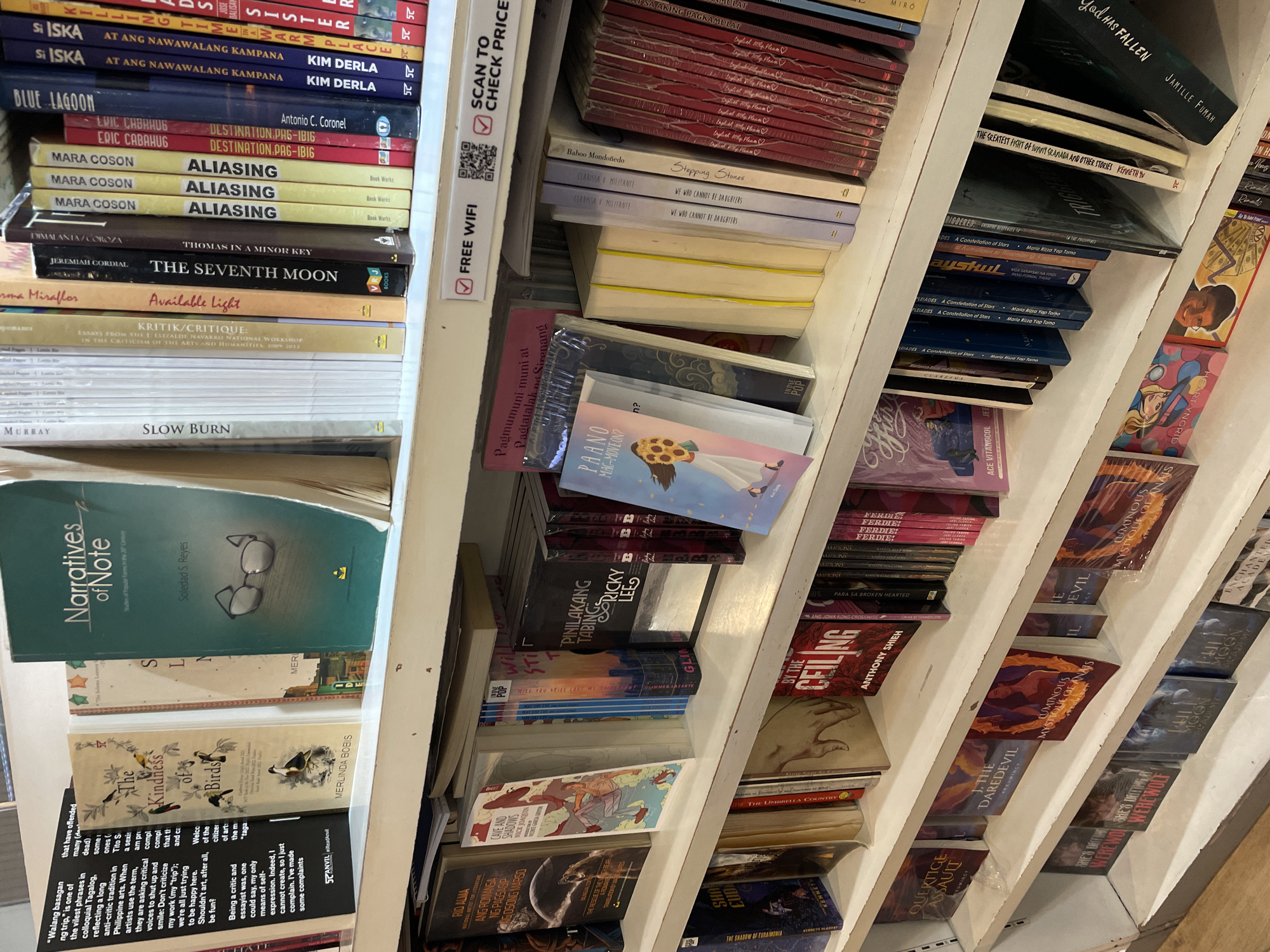 The image depicts a close-up, eye-level view of several bookshelves filled with books, likely within a bookstore or library. The shelves are white and appear to be made of particle board or a similar material. The books are of various sizes, colors, and bindings, with titles visible on many of their spines.
On the top shelf, prominently displayed, is a book titled "Narratives of Note" by Soledad S. Reyes, featuring an illustration of eyeglasses. To its left are books with titles such as "The Kindness of Birds" by Merlinda Bobis, and a book with text in Filipino. Further to the left, partially obscured, are books with text in English, including one with the title "Walang basagan ng trip."
The middle shelf showcases a diverse collection of books. Notable titles include "RIO ALMA," "ANG ROMANSA NG PAGSASANG OSANG MAPSO," "CAVE AND SHADOWS," "PINILAKANG TABING," and "PAANO MAG-MOVE ON?". A sign above this shelf indicates "FREE WIFI" and a QR code for "SCAN TO CHECK PRICE."
The bottom shelf displays more books, some with eye-catching cover art. Titles include "OVERTURE ASSAULT," "J. THE DAREDEVIL," "LUMINOUS MYSTIQUE OF ANAIS," and "SHE'S DRIVING A WEREWOLF."
The lighting appears to be artificial, casting a uniform glow on the books. The overall environment suggests a retail or public space dedicated to books.
Based on the inclusion of titles in Filipino and the reference to "Davao City, Philippines" in the prompt, this scene is likely situated in a bookstore or library within Davao City, Philippines. The presence of Filipino language titles on books like "Walang basagan ng trip" and "PAANO MAG-MOVE ON?" strongly supports this geographical context.