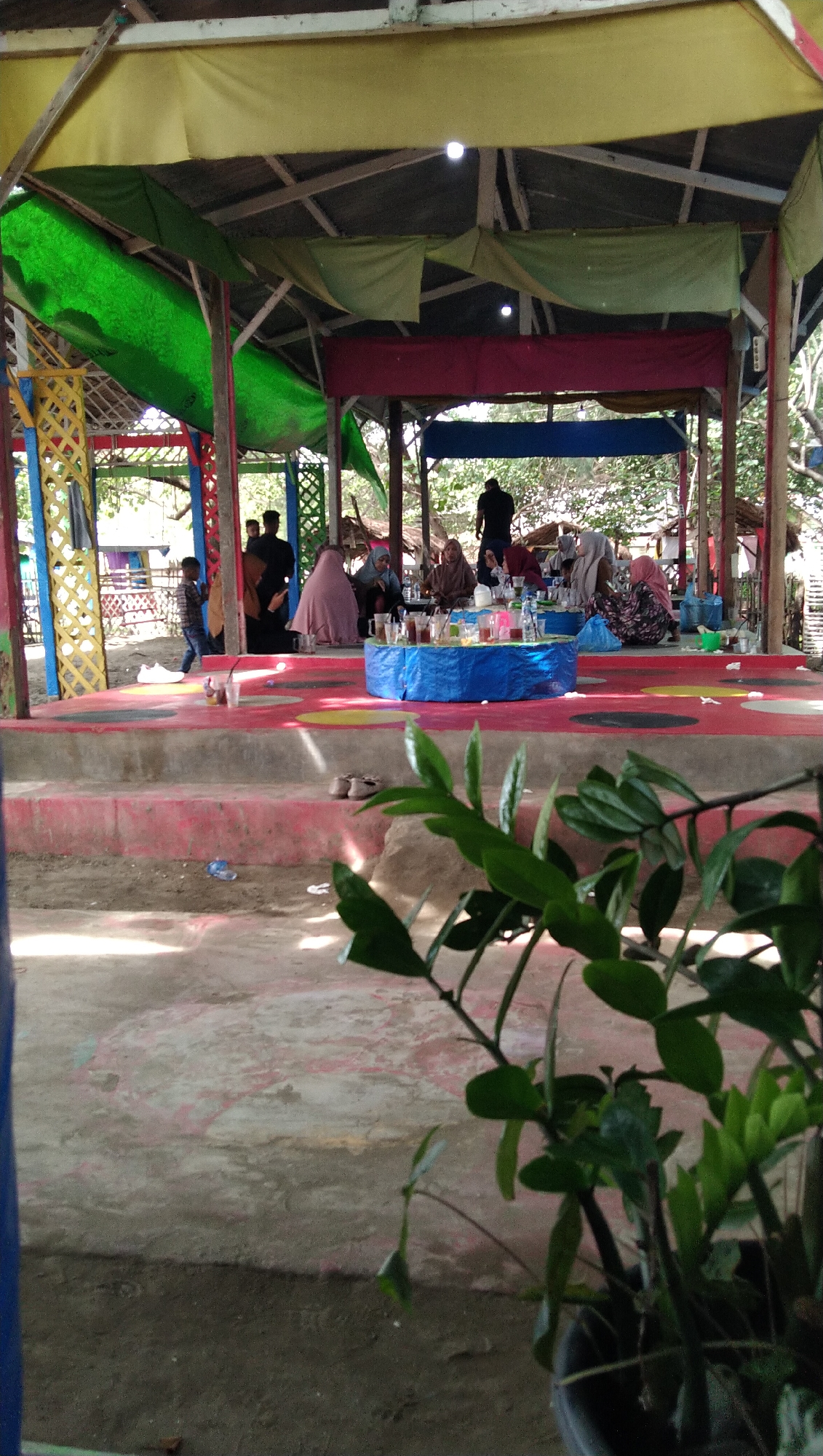 A group of people, some wearing hijabs, are gathered around a round, blue-covered table. They appear to be sitting on the floor in an open-air pavilion with a multi-colored fabric roof. Several bottles of drinks and cups are visible on the table. In the foreground, a potted plant with green leaves partially obscures the view. The ground in front of the pavilion is concrete with a red and pink painted surface. The pavilion is supported by wooden pillars and decorated with brightly colored elements. In the background, trees and more rustic structures are visible, suggesting an outdoor recreational area. The overall scene depicts a casual gathering or meal.