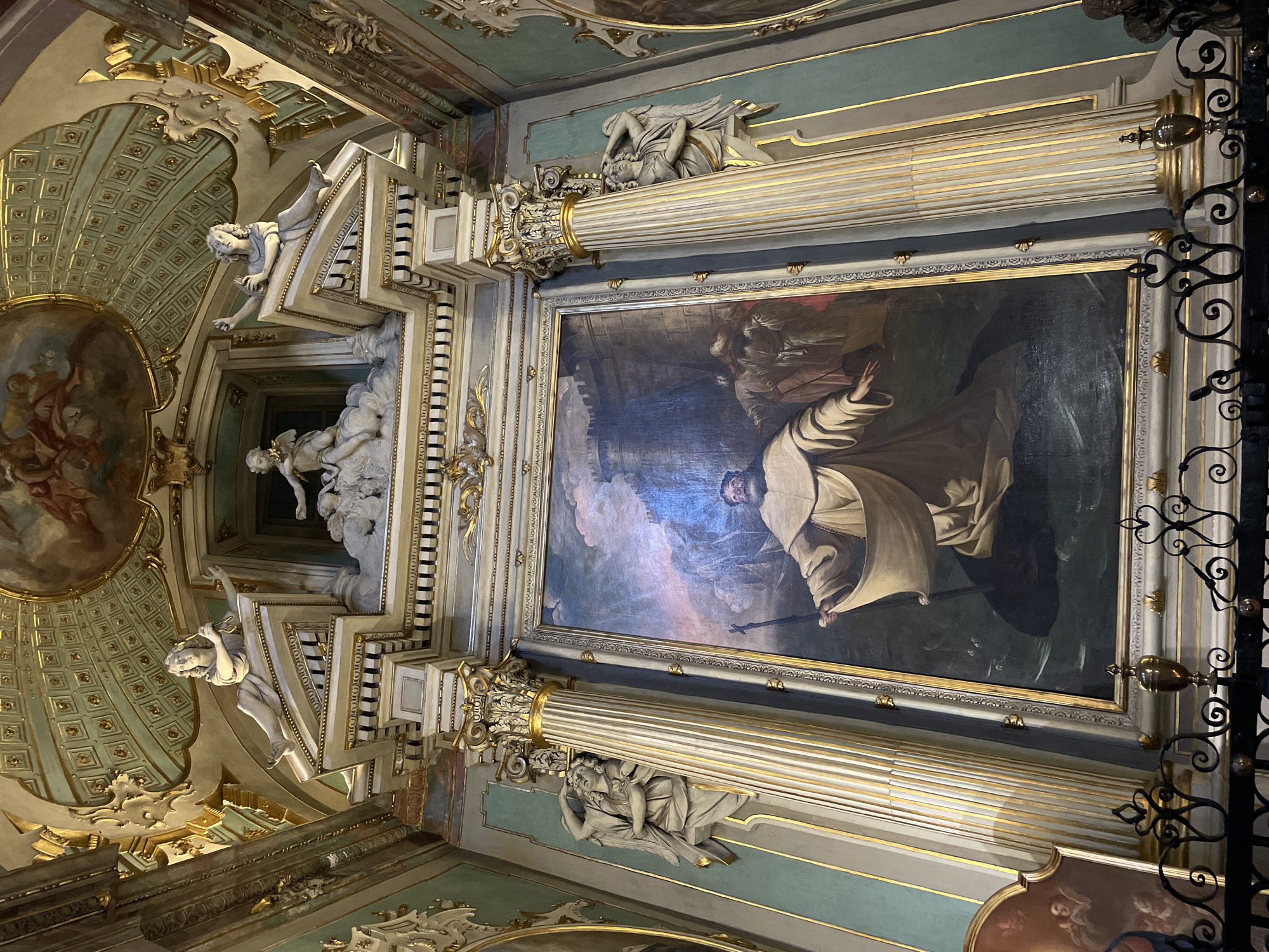 This image captures an ornate ceiling in a room in Bergamo, Italy. The ceiling features a large rectangular painting framed by decorative columns. The painting depicts a man in a white robe with his arm outstretched. Other figures appear in the background of the painting. The columns supporting the painting are decorated with sculpted figures and gold accents. The surrounding ceiling has intricate patterns, architectural details, and additional sculpted figures. There are also sections of painted frescoes.