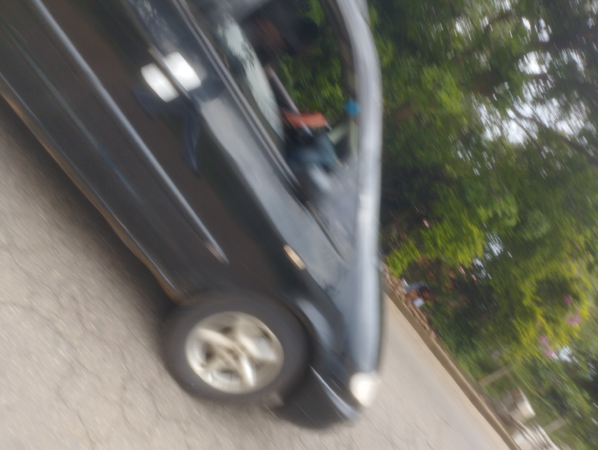 The image shows a side view of a dark-colored car moving along a paved surface. The car's wheels are visible, with the front wheel on the right side of the frame showing a silver rim. The car appears to be in motion, as indicated by the blurriness of the image, particularly the background foliage and the road surface. Through the car windows, it is possible to discern the blurred shapes of people inside. The environment appears to be outdoors, with lush green trees and vegetation visible in the background, suggesting a location with significant greenery. The lighting conditions indicate it is daytime, with some bright reflections on the car's surface. The scene is consistent with a street or road in a somewhat developed urban or semi-urban area. Without further information or context, it is not possible to definitively confirm the location as Jos, Nigeria, although the presence of trees and paved roads is common in such locations.
