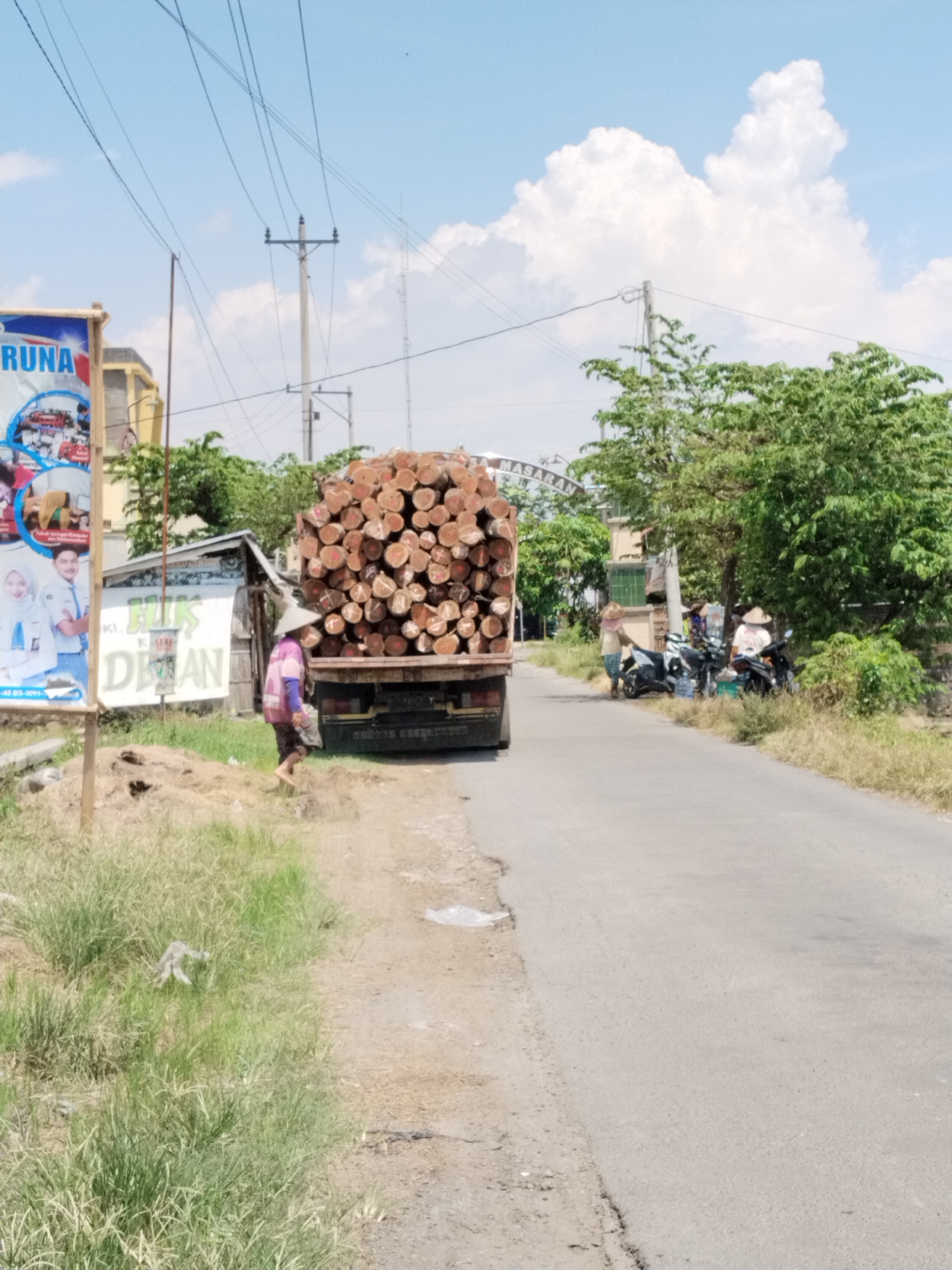 This photograph captures a sunny, midday scene on a rural or semi-rural road in Masaran, Indonesia. A large, dark-colored truck, heavily laden with numerous neatly stacked, light-brown cut logs, dominates the center-left of the image. The logs are piled high, showcasing their cross-sections.
On the left side of the road, a person wearing a conical hat and light clothing, possibly a pink shirt, walks barefoot on the grassy dirt shoulder, heading away from the camera and towards the front of the truck. Adjacent to this, a large billboard is partially visible, featuring the text "RUNA" and images of young people, including students in uniforms, suggesting an educational or promotional advertisement. Behind the walking person and next to the truck, there's a small, makeshift roadside structure with a white banner displaying green text, "JELIS" and "SDAN" being partially visible.
Further down the road on the right, several motorcycles are parked, and two or three individuals, also wearing conical hats, are gathered around what appears to be another small roadside stall, possibly selling drinks as bottles are visible. The road itself is asphalt, showing some cracks along its edges, and is flanked by patches of tall grass and lush green trees and bushes under a bright blue sky dotted with fluffy white clouds. In the distance, an archway can be seen, clearly bearing the text "MASARAN", confirming the location. Utility poles with power lines crisscross the sky, adding to the rural landscape.