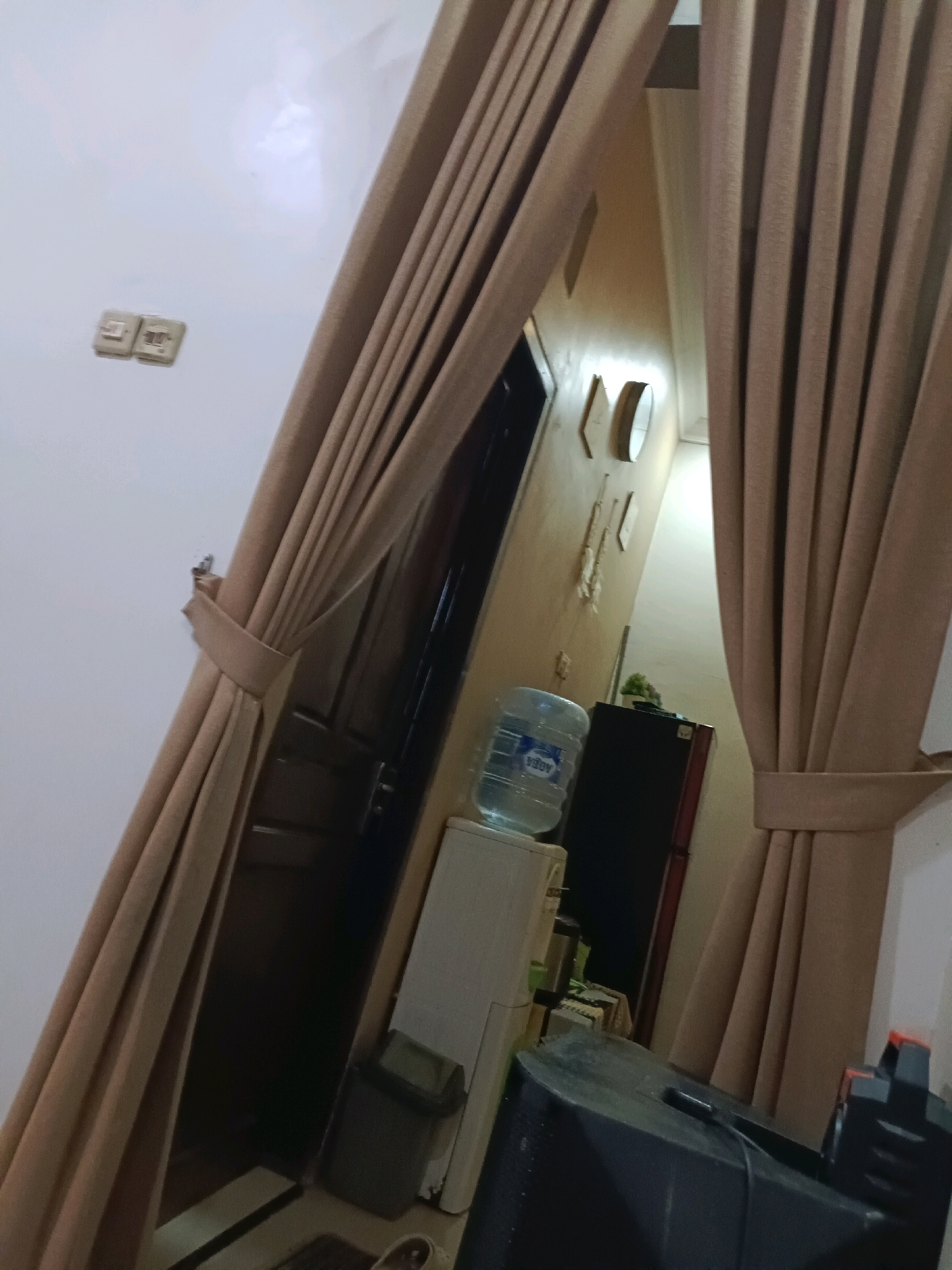 The image shows an interior scene, possibly a hallway or entrance area, in Rancang Kencono, Indonesia. The dominant features are large, beige curtains that partially obscure the view of the space. To the left, on a white wall, are two light switches. Further into the scene, a doorway is visible, leading to a darker area. Next to the doorway, on top of a white appliance, is a large blue water bottle. To the right of the water bottle, a dark-colored refrigerator is present. A portion of a black object, possibly a piece of furniture or electronic equipment, is visible in the foreground on the right. The lighting appears to be dim, contributing to the shadowed areas within the space. The overall impression is of a functional, perhaps lived-in, domestic environment.
