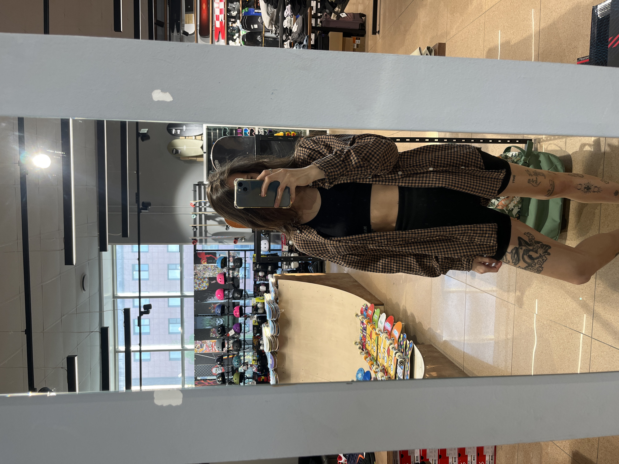A person is taking a mirror selfie in a store, likely in Krasnoyarsk, Russia. The person is wearing a black top, black shorts, and a brown checkered shirt. They are holding a smartphone in their hand to take the picture. The mirror reflects the store's interior, which includes rows of skateboards, helmets, and other sporting goods. A ramp is also visible, indicating the store is likely a skate shop. The store has large windows that reveal a building across the street. The floor appears to be tiled.