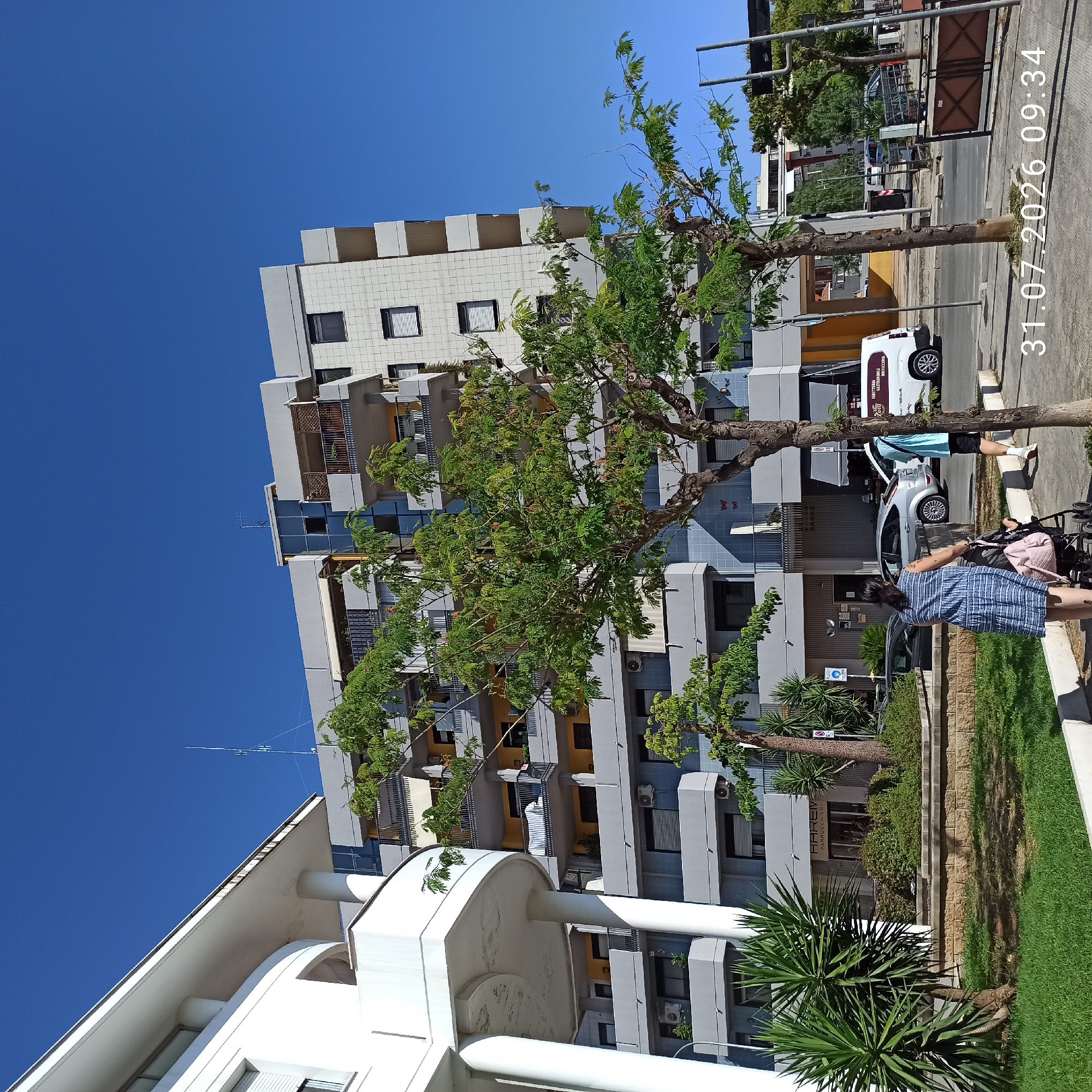 The image shows a street scene in Modugno, Italy, with a modern apartment building dominating the background. The building features multiple balconies with railings and is painted in shades of gray and white. In the foreground, a woman in a blue and white striped dress pushes a stroller, and a person in a light blue t-shirt and black shorts walks away from the camera. Trees with green foliage are present on the sidewalk and in front of the building. A white van with text on its side is parked on the street. The sky is clear and blue. The date and time "31.07.2026 09:34" are superimposed on the lower right corner of the image.