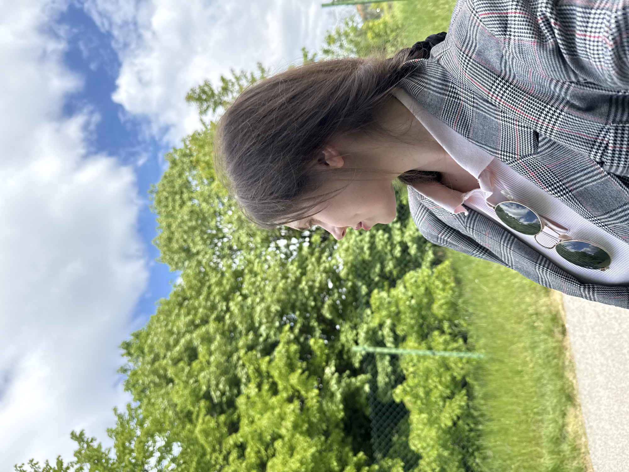 A young woman is captured from a low-angle perspective, seemingly looking down. She has brown hair styled with a braid visible at the back. She is wearing a gray plaid blazer over a light pink top with a bow detail at the collar. A pair of sunglasses is tucked into the blazer's lapel.
The background consists of lush green trees and a bright blue sky with scattered white clouds. A portion of a path or road is visible in the lower right corner of the image.
Without explicit landmarks, definitively identifying the location within Sremska Kamenica, Serbia, is impossible. Based on the natural environment, it may be a park area within the city.