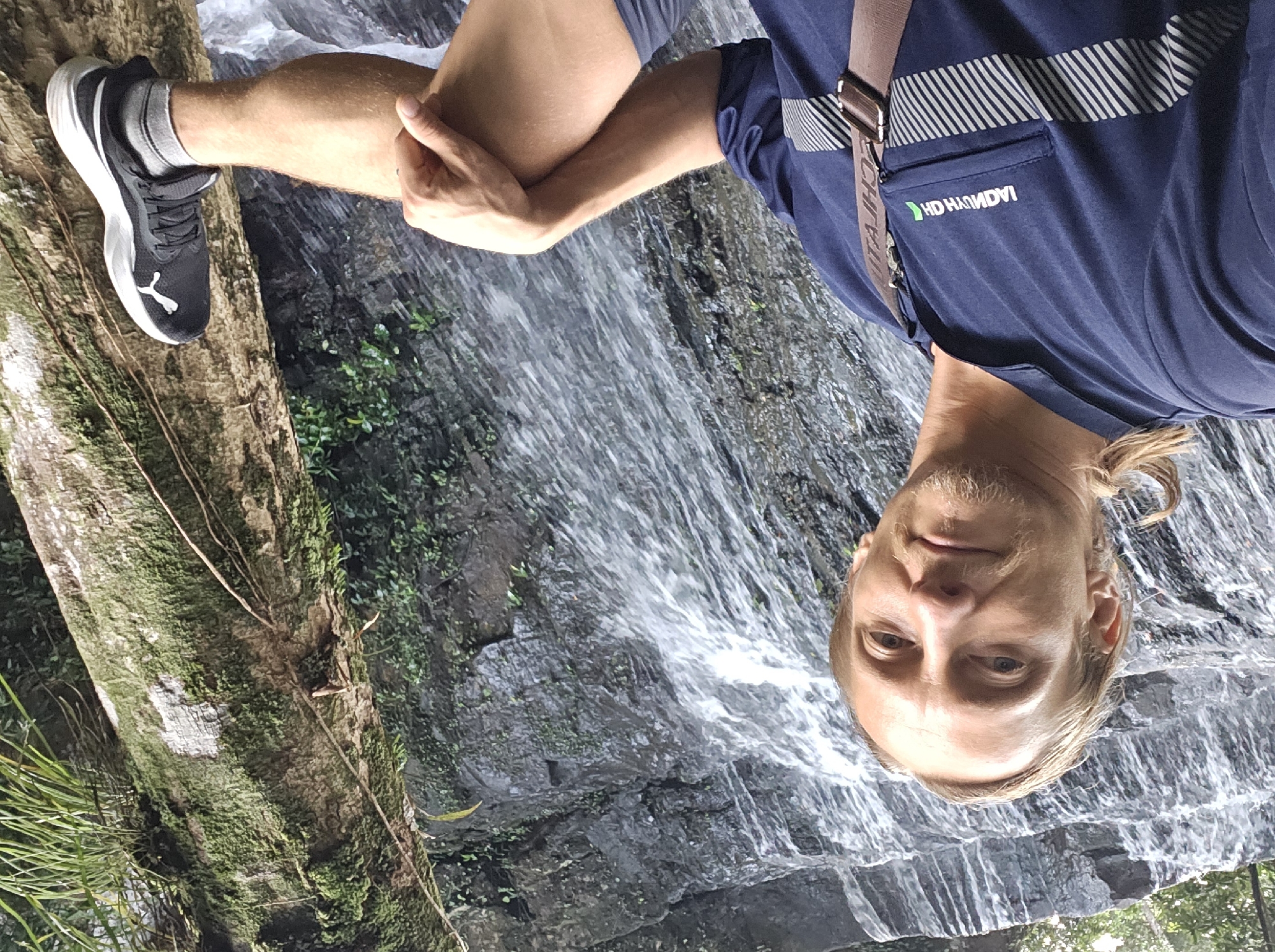 In Sok San Village, Cambodia, a light-skinned male with blonde hair tied back and a goatee is pictured from the chest up, leaning against a tree with his right leg extended. He is wearing a dark blue polo shirt with a silver striped pattern near the hem and a brown strap across his chest. A portion of a logo is visible on his shirt, reading "HD HANDY" with a green symbol to its right, and an inverted "TTACHJAI" is seen on the strap. His right hand rests on his knee, and his right foot, clad in a black and white Puma athletic shoe with grey socks, is placed on the tree. Behind him, a waterfall cascades down a rocky, moss-covered cliff face, surrounded by lush green foliage. The tree he is leaning against is also covered in moss and vines.