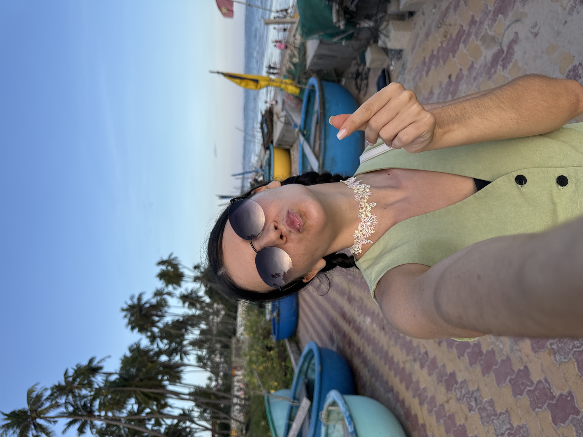 A woman is taking a selfie on a beachfront promenade in Mũi Né, Vietnam. She is wearing a light green sleeveless vest over a black top, a white lace choker, and large sunglasses. Her lips are puckered, and her right hand is making a heart shape gesture. She has dark hair styled in two braids. Behind her, several round, shallow boats, typical of Vietnamese fishing villages, are lined up on the promenade. Further in the background, palm trees line the coast, and the sea is visible with a few people on the beach and some fishing nets. The ground is paved with patterned tiles. The sky is clear and blue with a hint of sunset colors.