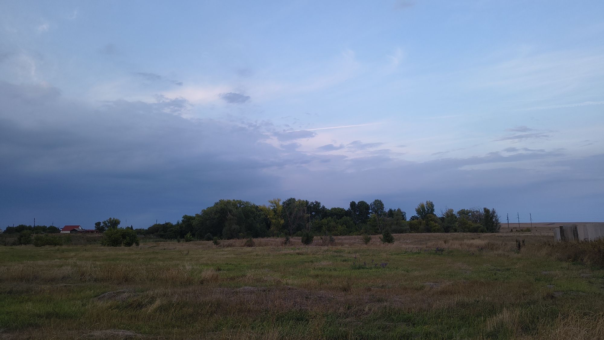 The image depicts a rural landscape at dusk, likely in the vicinity of Sharapensky, Russia. The sky is a gradient of blue and grey, with scattered clouds and a faint contrail from an aircraft. In the foreground, a dry, grassy field stretches across the frame. A line of trees forms a dark silhouette against the horizon, with some houses visible to the left, one with a red roof. To the far right, a concrete structure and utility poles are visible in the distance, suggesting the edge of agricultural land or a sparsely populated area. The overall scene conveys a sense of quietude and the transition from day to night. There are no people or animals visible in the image.