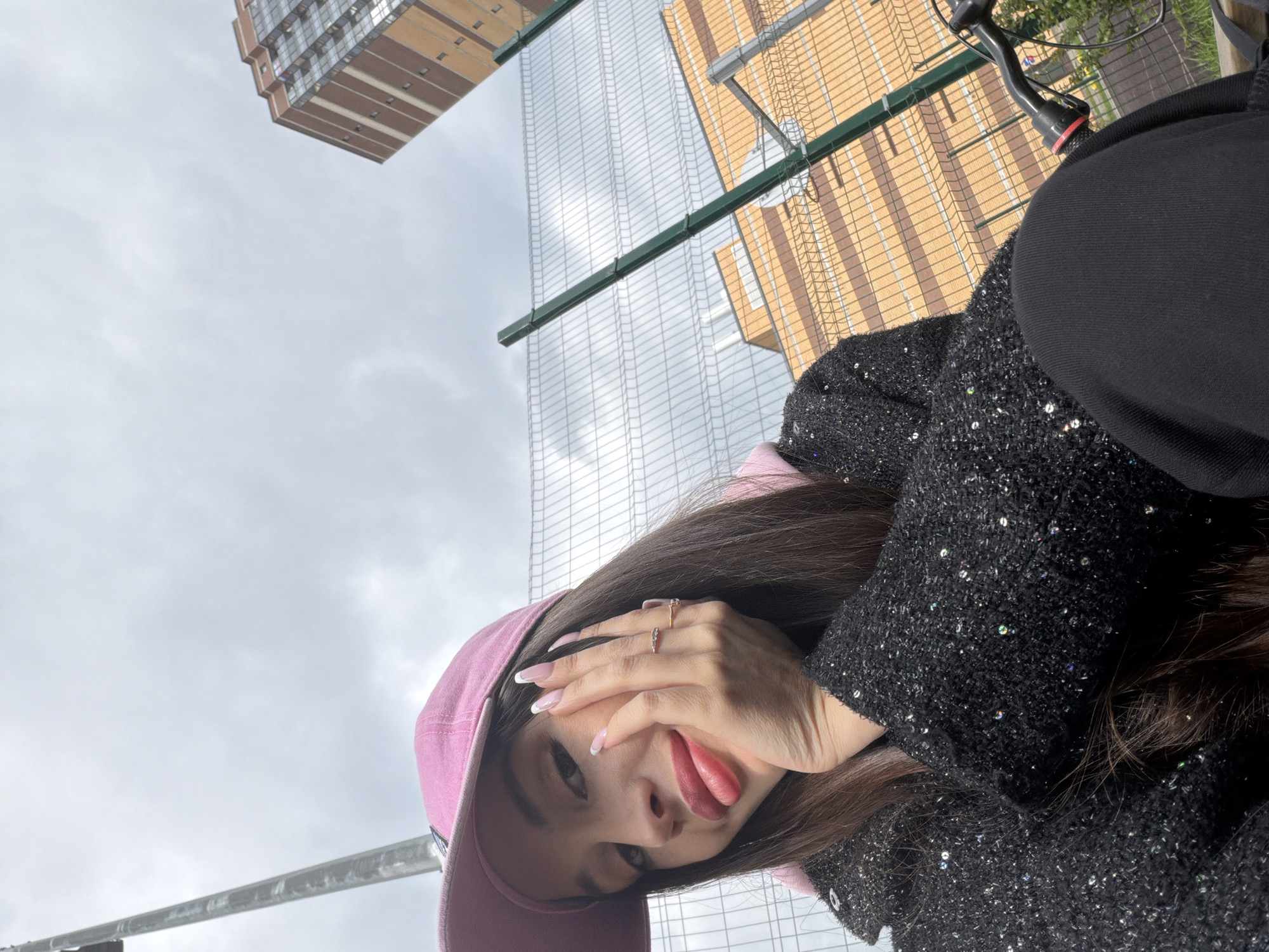 A woman with long, dark hair wearing a pink cap and a black, sequined jacket is posing with her tongue out. She is wearing two rings on her fingers. The background includes the sky and the tops of buildings, including a building with brown brick and a glass-covered skyscraper. The image appears to be taken outdoors. The location is identified as being in the city of Saint Petersburg, Russia.