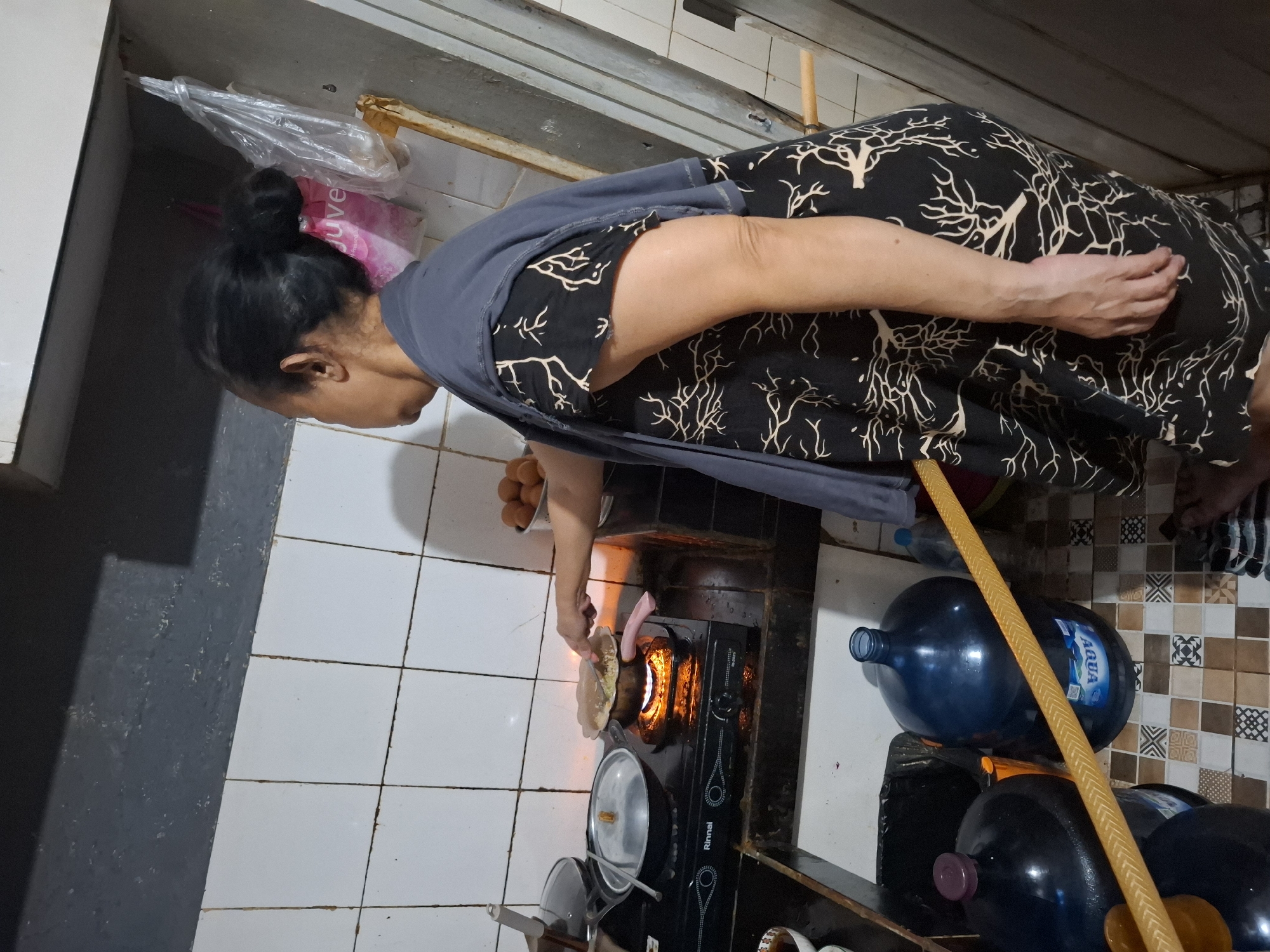 The image depicts an indoor scene, likely a kitchen, in West Jakarta, Indonesia. A woman with her hair pulled up in a bun is standing in front of a gas stove, actively cooking. She is wearing a dark dress with a light-colored tree-like pattern and a grey shawl draped over her shoulders. The stove has a flame visible from one of the burners, indicating that food is being cooked in a pot or pan. To the left of the stove, there are two covered pots. On the right side of the stove, a bucket of eggs is visible. The kitchen walls are tiled with white squares. In the foreground, several large blue water jugs are present. A yellow hose or tube is lying across the water jugs. The overall environment appears functional and used.