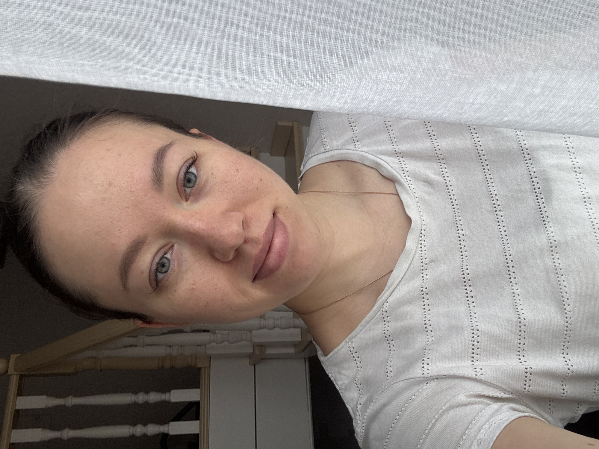 A woman with fair skin and blue-green eyes is photographed indoors. She is facing the camera with her head slightly tilted to the right. Her hair is dark and pulled back. She is wearing a white shirt with small, evenly spaced vertical stripes of tiny holes. A thin gold necklace is visible around her neck. A white, semi-transparent curtain hangs above and partially behind her. A section of what appears to be light-colored wooden furniture can be seen in the lower left corner of the frame. The location is Ферма, Russia.