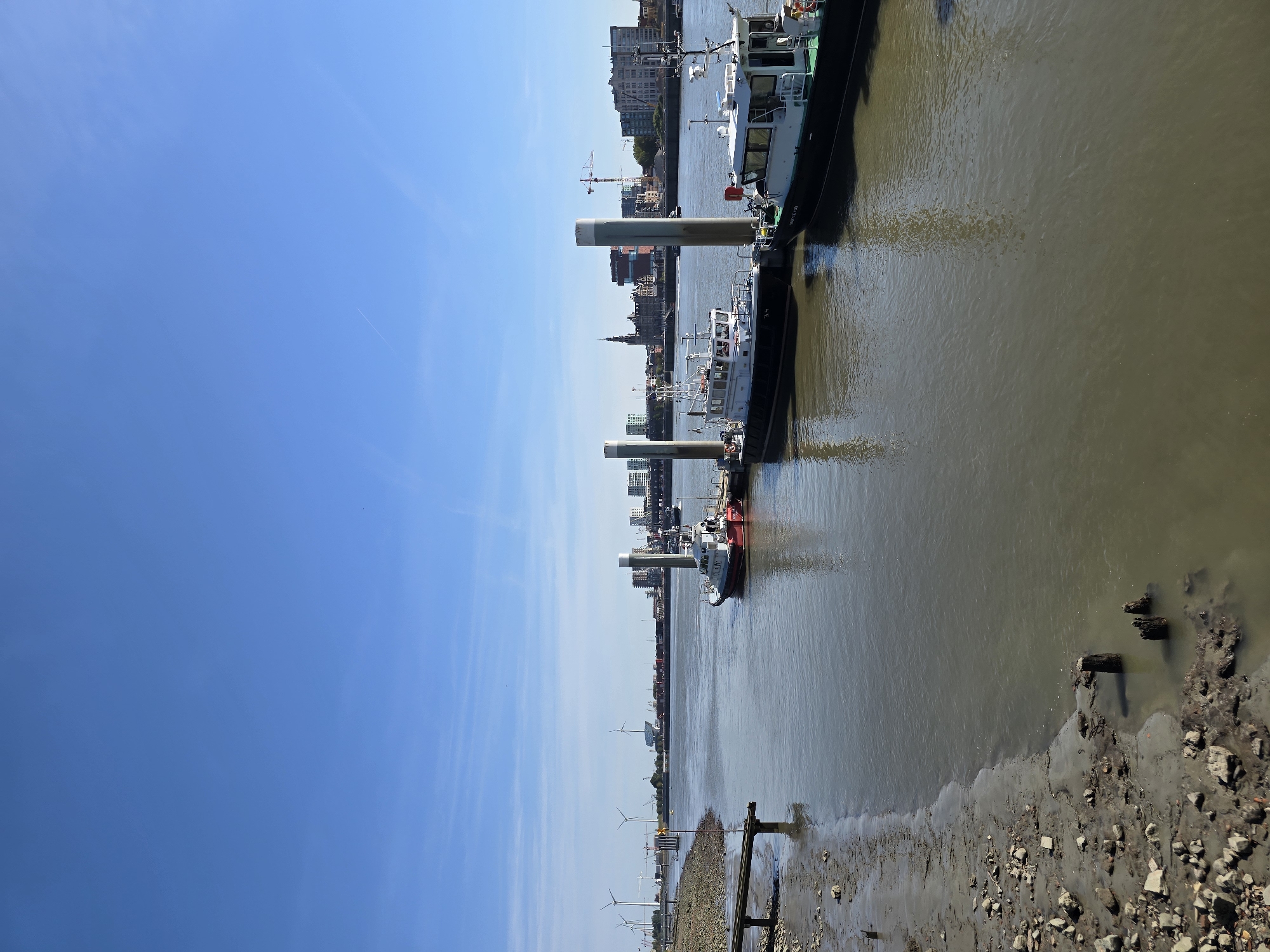 This is a wide-angle, landscape photograph of the Scheldt river in Antwerp, Belgium, on a clear, sunny day. The foreground features a muddy, rocky shoreline with several wooden pilings partially submerged in the water. In the mid-ground, a wide, calm river flows, reflecting the blue sky and distant structures.
Three boats are docked along a modern pier with white cylindrical bollards. The boat closest to the foreground on the right is dark green and white, with the text "ZEEWERKER" visible on its side. To its left is a white boat with black trim, and further to the left is a smaller red and white boat.
The background displays the cityscape of Antwerp, characterized by a mix of modern buildings, a prominent church tower, and industrial elements like cranes and wind turbines visible on the horizon. The sky is a vibrant, cloudless blue, indicating fair weather. The overall scene conveys a sense of industrial activity and port life along the river.