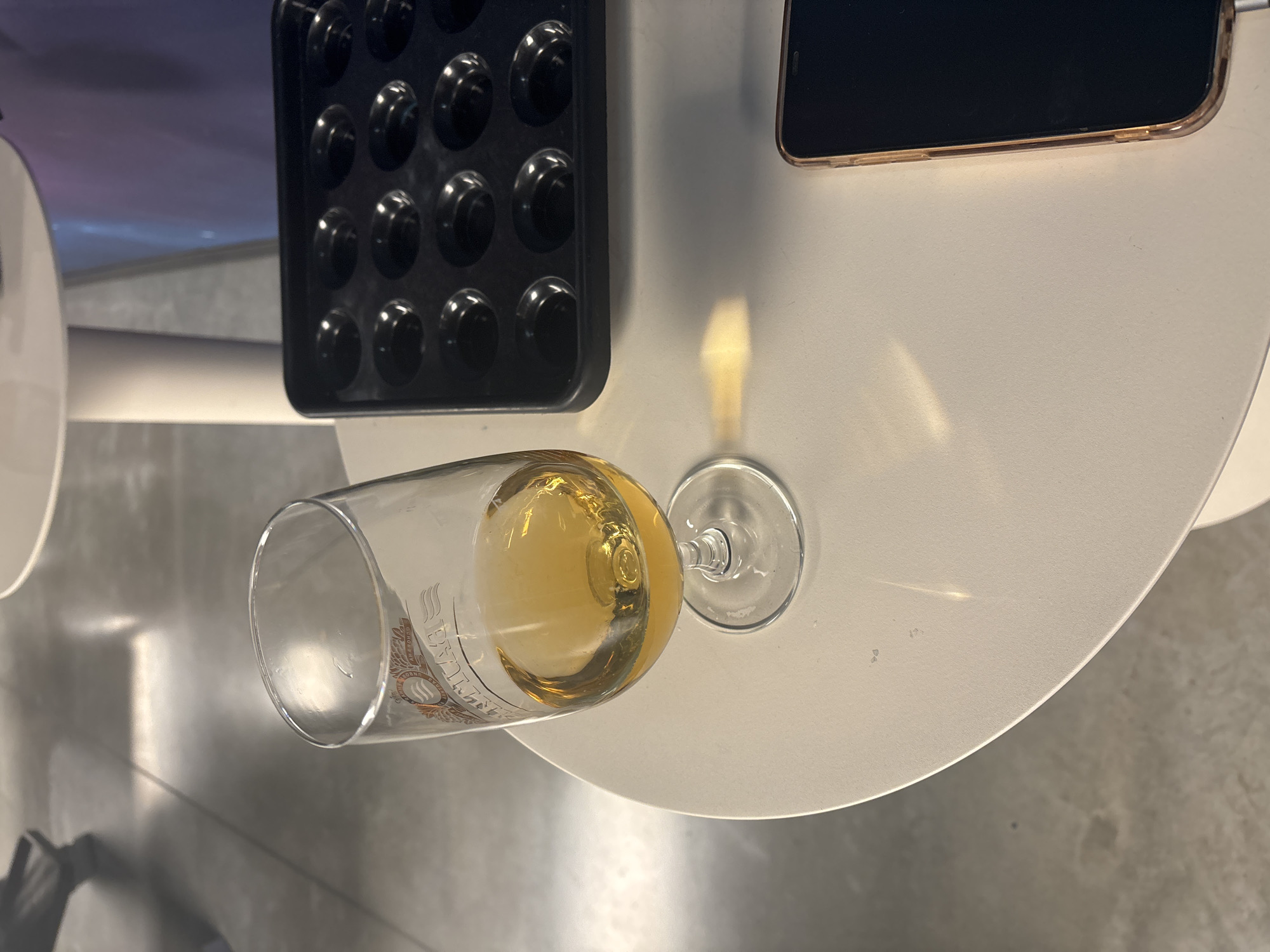 A close-up, high-angle shot focuses on a table with a glass of amber liquid and a black mold with circular indentations. The glass, positioned to the left, contains a golden-hued beverage, possibly whiskey or brandy, with a logo visible on its side. To the right of the glass is a black, non-stick baking mold, likely for mini muffins or chocolates, with twelve circular cavities. A sliver of a smartphone with a rose gold case is visible in the lower right corner. The table surface is white and appears to be made of a smooth material. The background is blurred, showing a grey, tiled floor and a portion of a white table or counter structure. The lighting suggests an indoor environment with a light source casting a reflection on the table. There are no people clearly visible in the frame, though shadows and blurred shapes in the background hint at their presence. The scene appears to be set in Saint Petersburg, Russia, possibly in a bar, cafe, or a home kitchen setting.