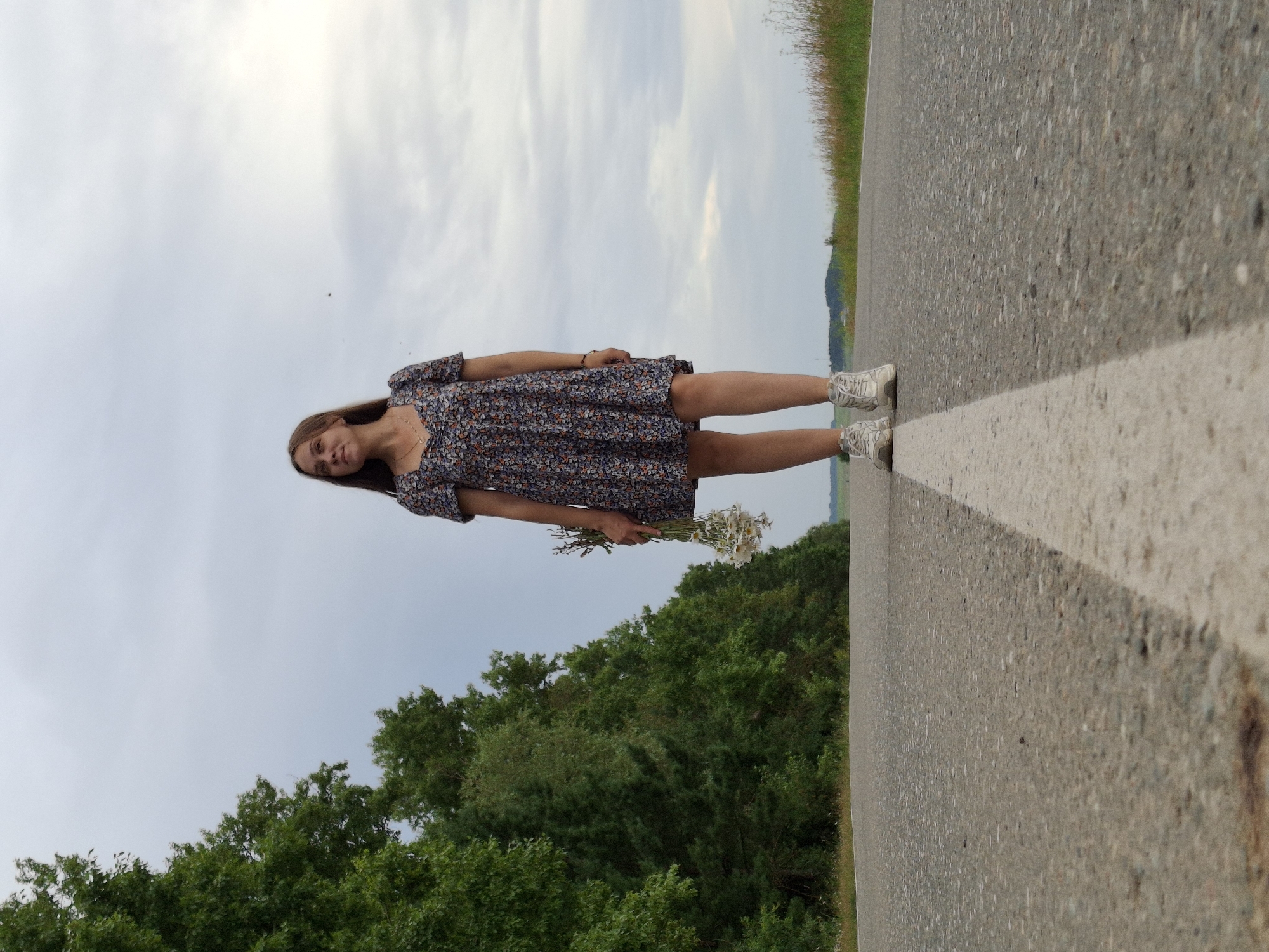 In a low-angle shot, a young woman stands on a paved road, facing slightly away from the camera towards the right. She is wearing a short, floral-patterned dress and white sneakers. A bouquet of white flowers is held in her left hand. The road is marked with a white line down the center, and it stretches into the distance. On the left side of the road, a dense line of green trees forms a backdrop. The sky above is overcast with a pale, cloudy texture. The scene appears to be in a rural or semi-rural setting, possibly on the outskirts of Гаревая, Russia, given the natural landscape and the presence of a road that could be an access route to or from the village. The overall mood is serene and contemplative.