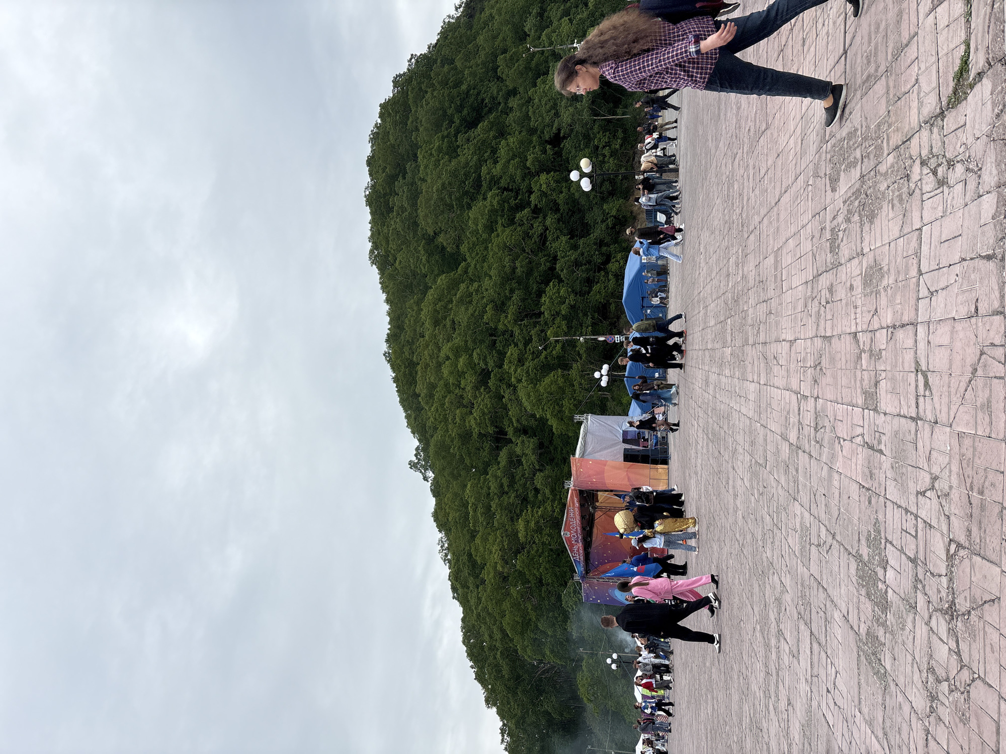 This image depicts a wide, paved plaza in Petropavlovsk-Kamchatsky, Russia, with a large, densely forested hill forming the backdrop. In the mid-ground, a stage with an orange and red backdrop displaying celestial imagery is set up, flanked by white tents. Several people are gathered around the stage, some in formal wear, others in casual attire, and one individual is wearing a gold costume. A woman with long, curly brown hair, wearing a plaid shirt and jeans, walks towards the right foreground of the image, in profile. The sky is overcast and cloudy. The plaza is paved with reddish-brown bricks, showing signs of wear and tear. Several lampposts with multiple bulbs are visible throughout the plaza.