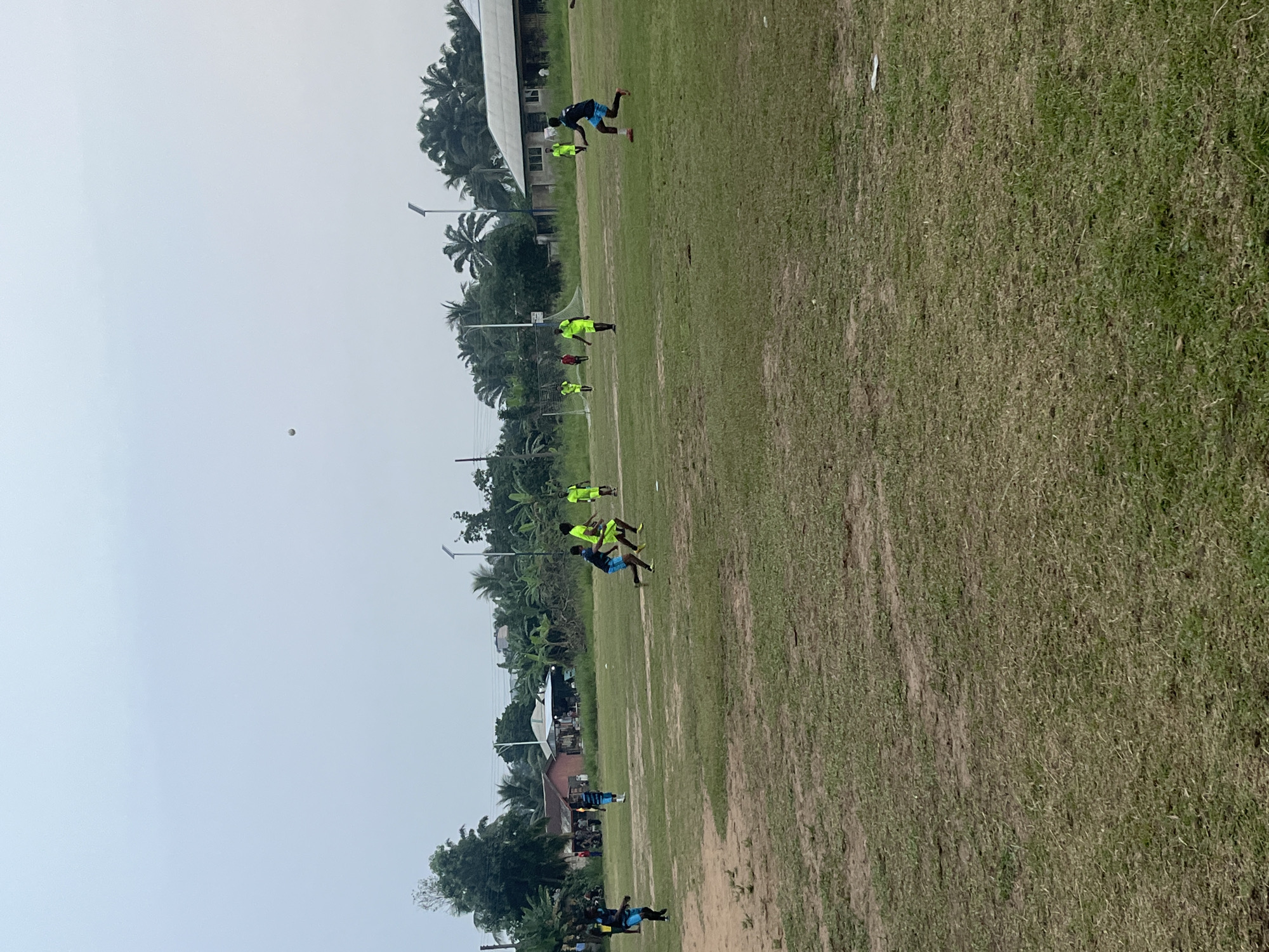 The image depicts a soccer game in progress on a grass field. Several players wearing neon yellow jerseys and a few in blue and black jerseys are seen engaged in the game. A white ball is visible in the air. The surrounding environment features palm trees, buildings, and a cloudy sky. The location is Unknown, Unknown.