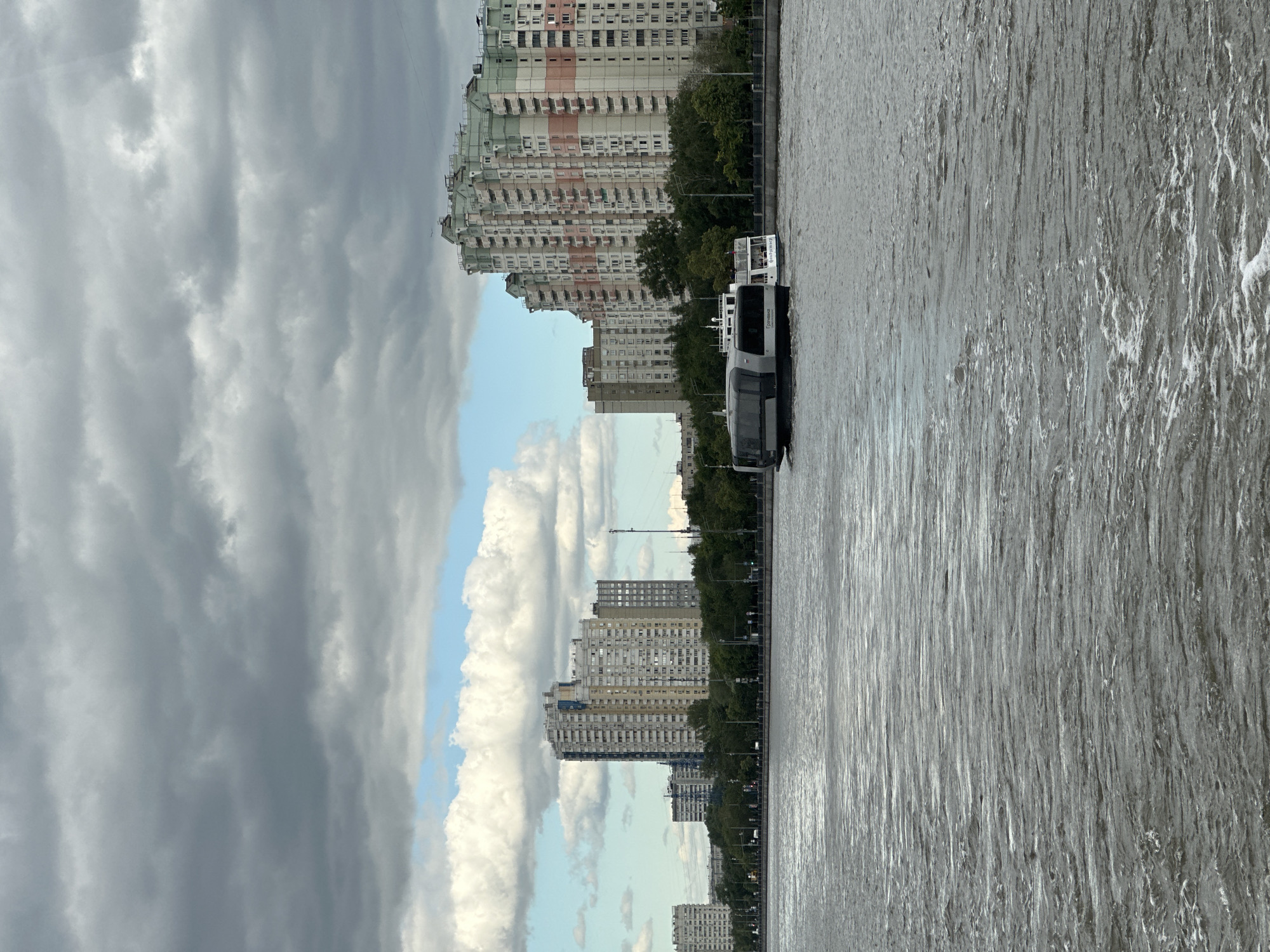 The image captures a view of the Moscow River on a cloudy day. A modern, sleek river boat with a dark gray hull and white upper deck is navigating the choppy water. In the background, a line of apartment buildings with colorful facades and a green-topped one dominate the skyline, interspersed with trees. The water is turbulent with visible ripples and foam. The sky is filled with dramatic, dark clouds, contrasting with patches of lighter clouds and a hint of blue sky.