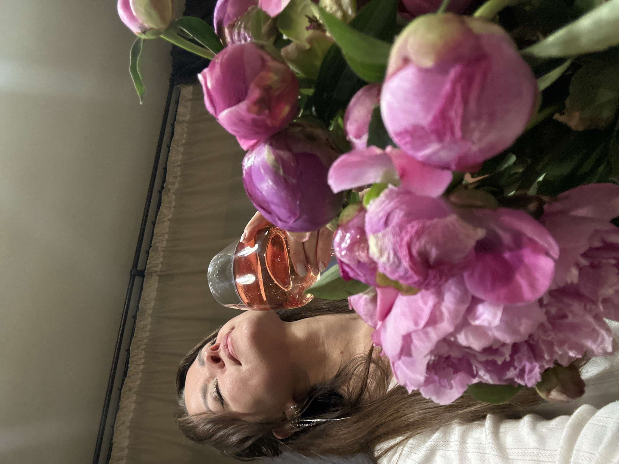 A woman with dark hair and closed eyes tilts her head back, holding a glass of rosé wine. She is wearing a white, ribbed top and a long, silver earring dangles from her left ear. In the foreground, a vibrant bouquet of pink and purple peonies fills the right side of the frame. The background appears to be an indoor setting, possibly a home, with a beige curtain and a dark ceiling visible. The lighting is warm and soft, highlighting the textures of the flowers and the woman's skin. The scene suggests a moment of relaxation and enjoyment.
