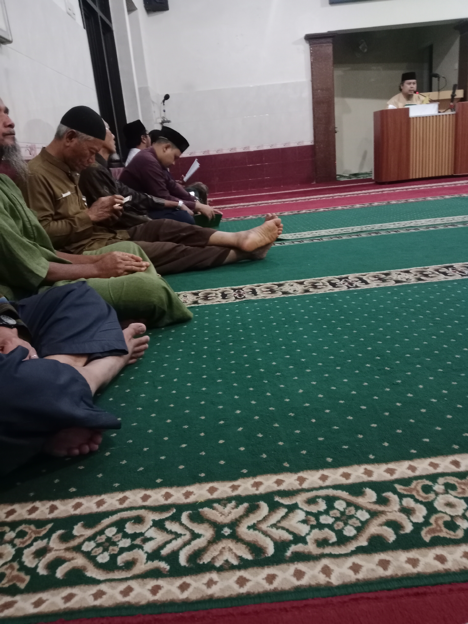 The image depicts an interior scene, likely a mosque, with several men seated on a green carpet with a decorative pattern. In the foreground, the legs and feet of at least two individuals are visible, one barefoot and wearing dark blue pants, the other wearing brown pants. Further back, men are seen sitting cross-legged, some wearing peci hats and traditional attire. One man in a brown garment is holding a mobile phone. In the background, a man in a light-colored robe is standing behind a wooden podium, appearing to address the seated congregation. The walls are white, with a section of dark red tiling near the floor. The overall setting suggests a religious gathering. This scene is consistent with the interior of a mosque in Kediri City, Indonesia, where such gatherings are common.