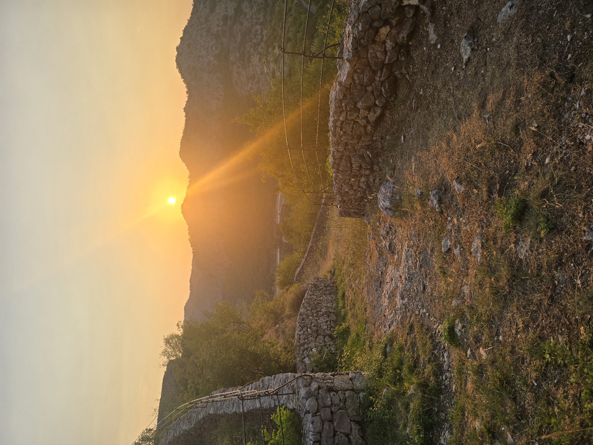 The image depicts a pathway lined with stone walls, leading upwards towards a mountain range bathed in the warm glow of a setting sun. The sky is a soft gradient of yellow and orange, with the sun just cresting a distant peak, casting long rays of light across the landscape. Green foliage and trees are scattered on the slopes of the mountains and along the sides of the path. The pathway itself appears to be made of dirt and gravel, with patches of grass and small stones. The stone walls, constructed from rough-hewn rocks, appear to be ancient and weathered. In the distance, the mountains are silhouetted against the vibrant sky. The scene suggests a rural, natural environment, likely in a mountainous region. The location is Gornja Vrbica, Montenegro.