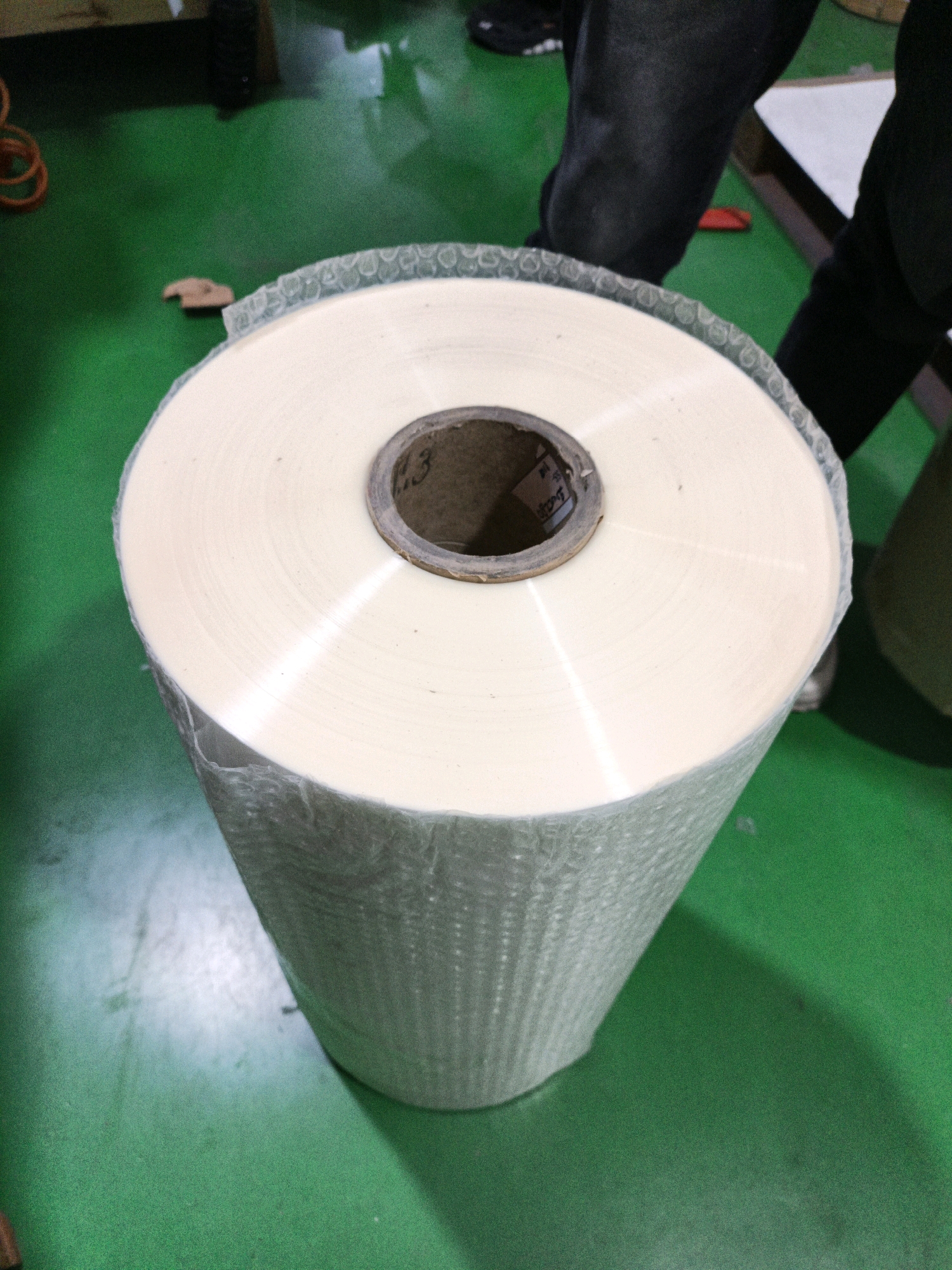 A large roll of white plastic film, wrapped in bubble wrap, stands on a green floor in what appears to be a factory or warehouse. The roll is centered in the frame, with its cardboard core visible and marked with some faint writing, including "1.3". The green floor has some scuff marks and there are parts of people's legs visible in the background, wearing dark clothing. To the left, a coiled orange cable is visible. The overall impression is of an industrial or commercial setting, possibly a manufacturing facility.
