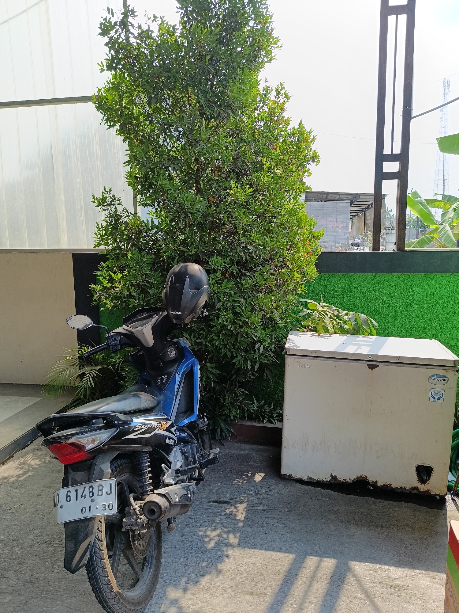 A blue and black motorcycle is parked on a concrete surface in front of a large green bush. A black helmet is resting on the handlebars of the motorcycle. To the right of the bush is a white chest freezer with rust on its bottom edge. In the background, a tall, dark metal structure is visible, along with buildings and trees under a bright sky. The scene appears to be an outdoor area in a city, likely Manisrenggo, Indonesia, given the license plate of the motorcycle which reads "AD. 6148 BJ 01-30".