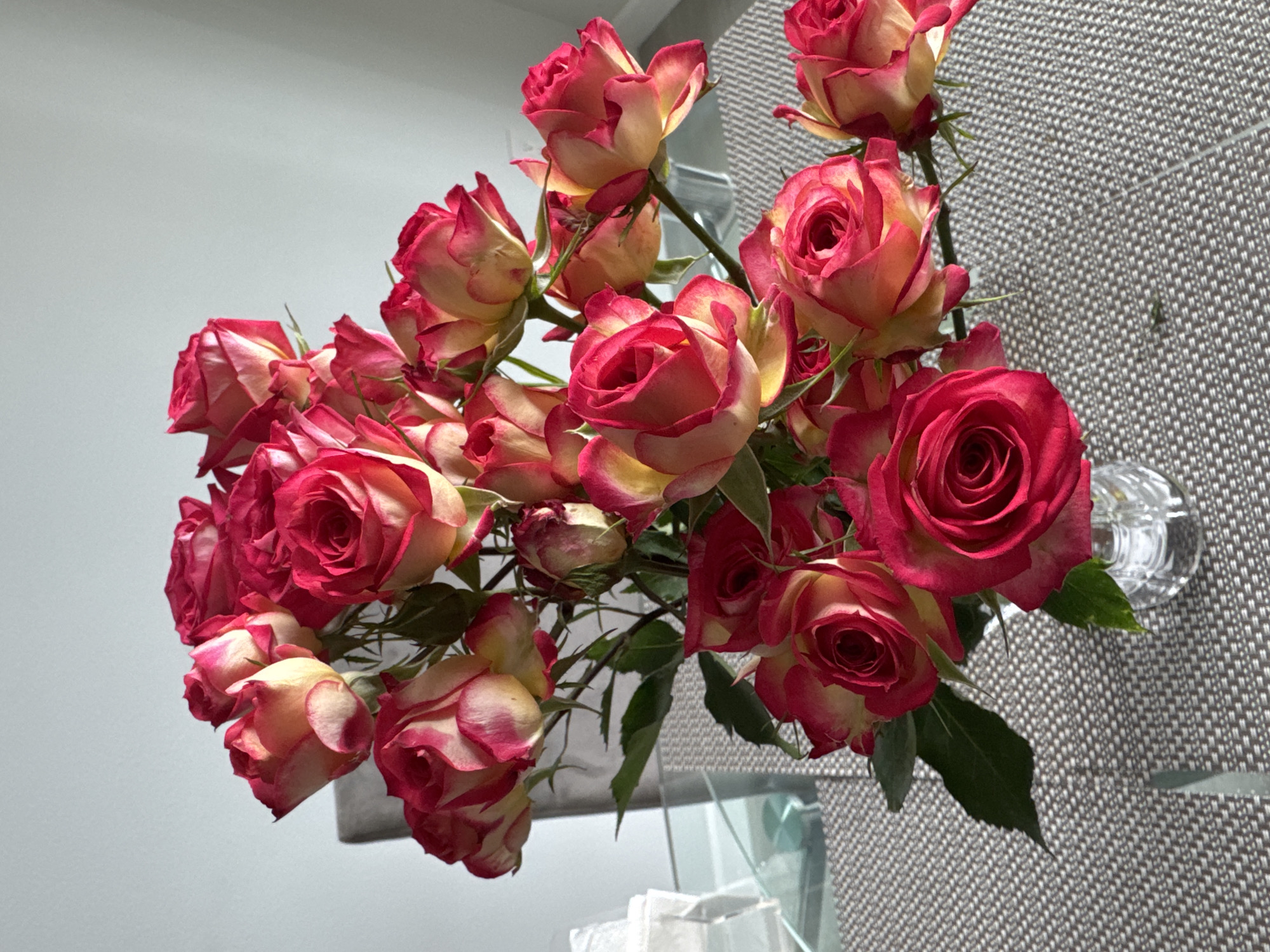 A bouquet of bi-colored roses, predominantly pink with yellow undertones, is displayed in a clear glass vase on a textured grey table. The roses vary in bloom stage, with some fully open and others still forming buds. Green leaves and stems are visible amongst the flowers. In the background, a light-colored wall and a sliver of a grey chair are present. The scene is lit by natural light, casting subtle shadows. The location is Viera, United States of America.