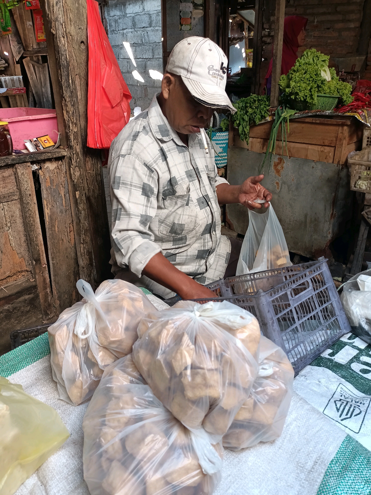 A man wearing a white baseball cap and a plaid shirt is sitting down in what appears to be an outdoor market stall in Badas, Indonesia. He is holding a cigarette in his mouth and is in the process of bagging some goods for a customer. In front of him are several plastic bags filled with what look like fried tofu or tempeh, resting on a white and green patterned cloth. To his right, a gray plastic crate is visible, and behind him, there are shelves stocked with various items, including a pink container, some jars, and a red plastic bag. Further in the background, a woman wearing a red headscarf is visible, along with a display of fresh green lettuce and red chilies. The stall appears to be made of rustic wooden materials, with a brick wall visible in the background. The overall scene depicts a transaction taking place at a local market.