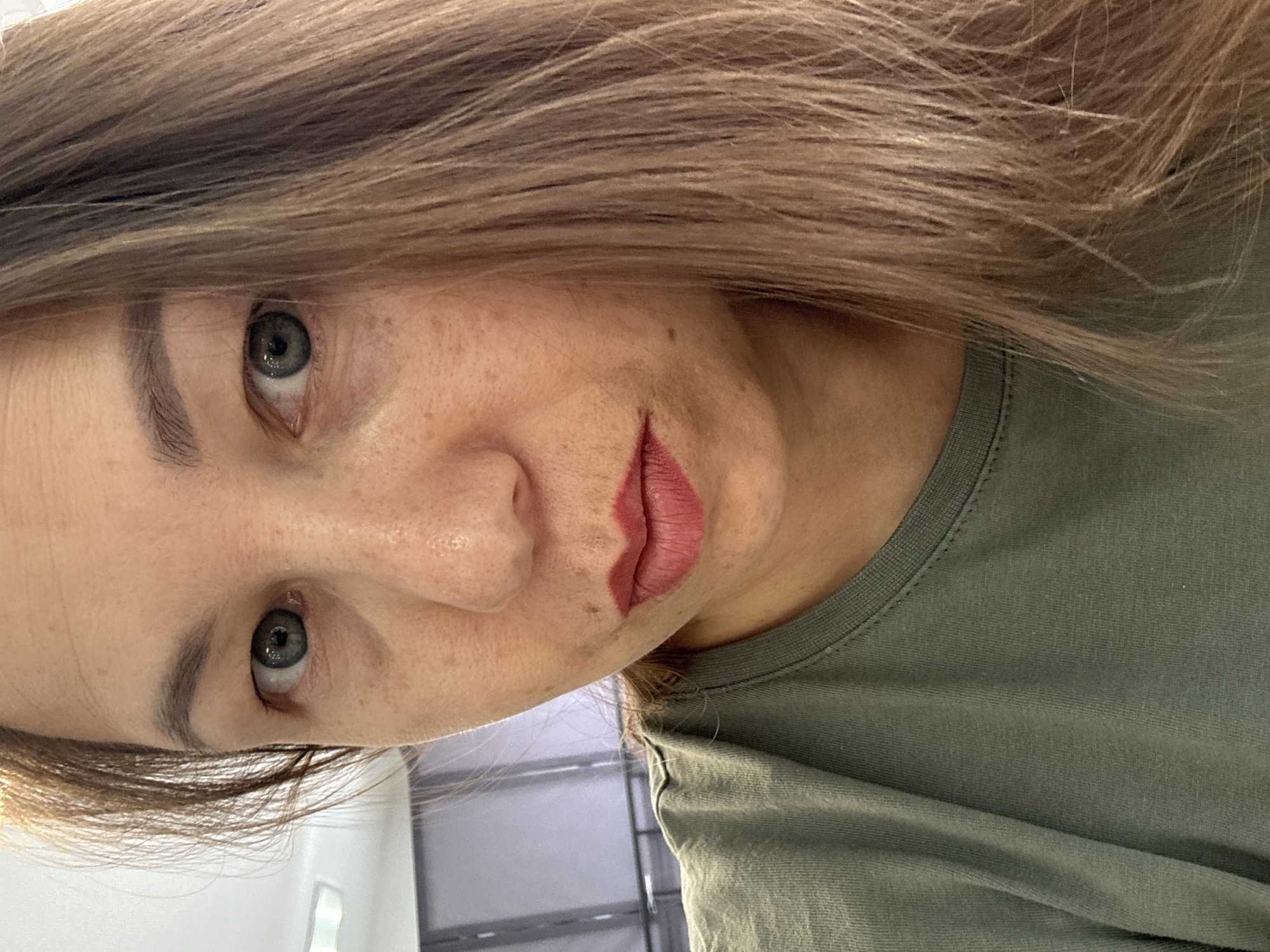 The image is a close-up, angled portrait of a person, likely female, captured from a slightly low perspective. Her head is tilted, and she gazes directly into the camera with a neutral to subtly contemplative expression. Her eyes are a striking grey-blue or greenish-grey. Her lips are painted with a red or deep pink lipstick and are slightly parted. Her skin displays natural texture and a scattering of freckles. Light brown or sandy blonde hair, appearing fine, frames her face and falls across her forehead. She is dressed in a plain olive green crew-neck t-shirt. The background is out of focus, suggesting an indoor setting with soft, diffused lighting, possibly from a window or artificial source, creating gentle highlights. While the context indicates the photo was taken in Chaikovsky, Russia, this specific location is not visually discernible within the image. There is no visible text.