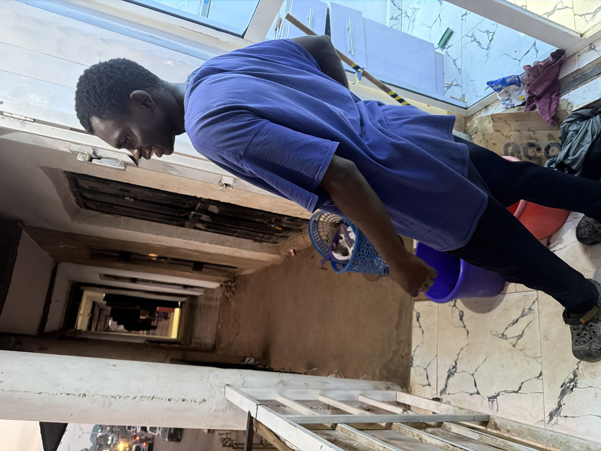 A young Black male is captured from a low angle, looking down into what appears to be a deep shaft or stairwell. He is wearing a purple t-shirt and black pants. His head is tilted down, and his expression is focused, suggesting he is observing something below. The background shows a tiled floor with a marbled pattern and some clutter, including a red bucket and a blue basket containing some white items.
The scene seems to be set in an interior, possibly a residential building or a communal space, with hints of external light suggesting it might be daytime. The overall atmosphere appears mundane, with the subject engaged in an activity that requires his attention. The depth of the shaft in front of him is emphasized by the diminishing perspective as it recedes into darkness, with glimpses of lights and structures far below. There is a broom with yellow and blue bands leaning against the wall behind him. A piece of white plastic sheeting or insulation is visible above and behind the man. The floor tiles are a light cream color with dark grey veining that resembles cracks. The wall behind the subject is a light tan color with some damage. There is a roll of what looks like toilet paper in a blue wrapper on a shelf to the right of the subject. Next to it, a dark pink cloth is draped over the shelf.
The image is taken in Bukuru, Nigeria.