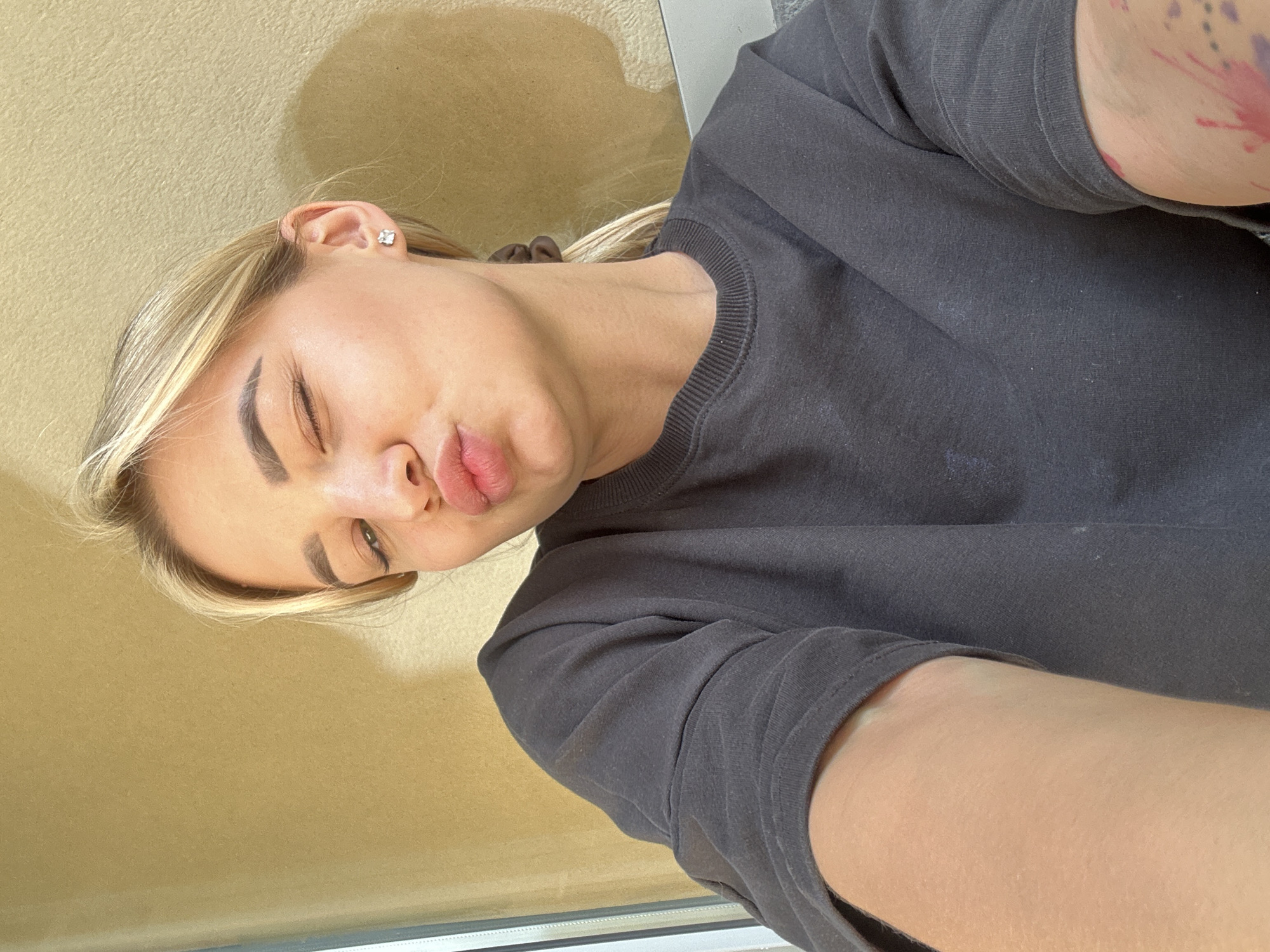 This close-up, rotated selfie features a young woman indoors. She has blonde hair, styled to the side, and is making a playful, pouty expression with her lips while her eyes are squinted or partially closed. She wears a dark grey crew-neck t-shirt or sweatshirt. A small, square, silver stud earring is visible in her right ear. On her upper right arm, partially covered by her sleeve, there is a visible tattoo featuring pink/red and purple colors. The background is a plain, light beige wall, with a shadow cast upon it, suggesting a strong light source, possibly sunlight from a window. A window frame or sill is visible at the very bottom of the image. The setting is an indoor location in Budva, Montenegro. No visible text is present.