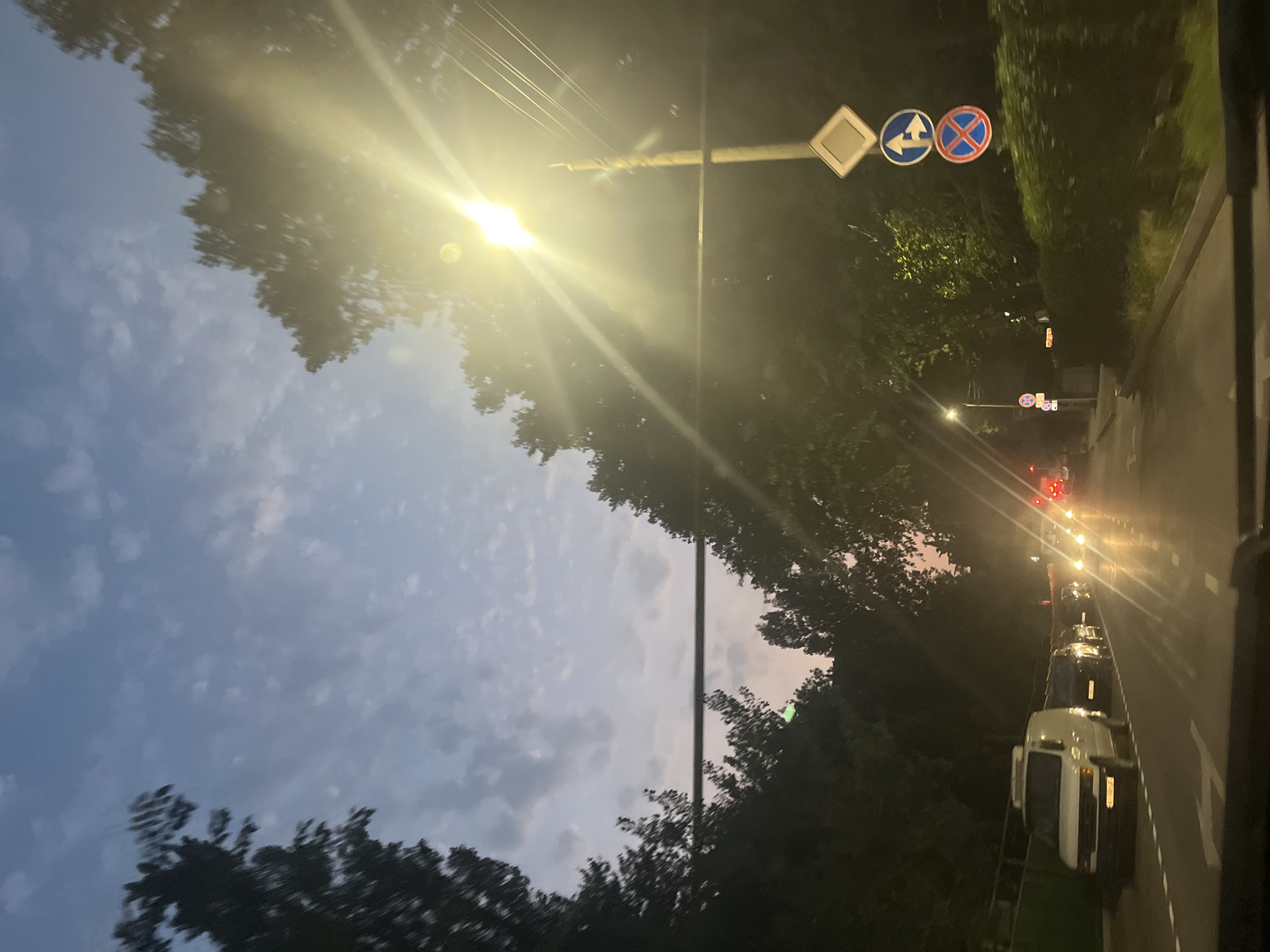 The image captures a twilight scene on a city street, possibly in Tbilisi, Georgia, with cars lined up in traffic. A bright streetlamp illuminates the right side of the frame, casting long rays and lens flares across the image. Trees line the sides of the road, their dark silhouettes contrasting with the dusky sky and the artificial light.
On the right side of the road, a lamppost displays three signs: a diamond-shaped sign at the top, followed by a circular blue sign with an arrow indicating a left turn and a straight direction, and a circular red and blue sign prohibiting stopping.
In the foreground, the view appears to be from inside a vehicle, with the dashboard partially visible at the bottom. A white van is prominent in the left lane, followed by several other cars. The road surface reflects the lights of the vehicles and the streetlamp, creating a streaky, wet appearance. The sky above transitions from a pale blue with scattered clouds near the horizon to a darker, purplish hue at the top, suggesting the sun has recently set. The overall atmosphere is one of a typical urban evening commute.