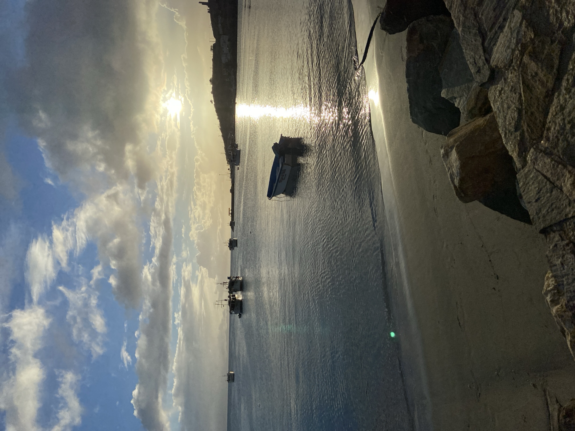 The image depicts a serene coastal scene at sunset in Juan Griego, Venezuela. The sun, partially obscured by clouds, casts a golden glow across the water, creating a shimmering path of light. Several boats are anchored in the calm bay, their silhouettes dark against the bright sky. In the foreground, a small, empty motorboat drifts gently near the shore. The beach is composed of dark sand and is lined with large rocks. The distant coastline is visible, with buildings and structures dotting the hillside, and a lighthouse-like structure can be seen on the right. The sky is a mix of blue and white clouds, with dramatic lighting from the setting sun.