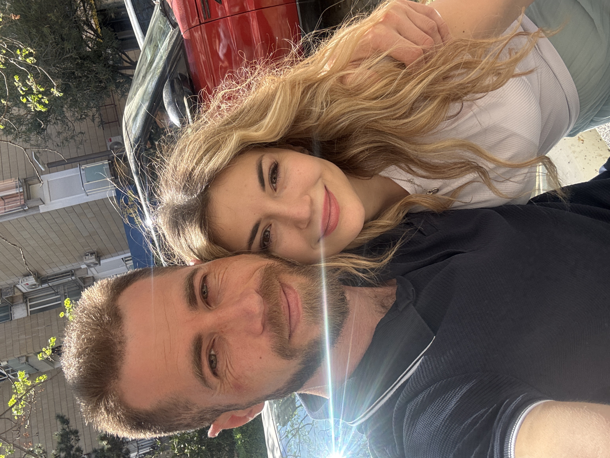 This close-up selfie, presented in a rotated vertical orientation, features a smiling couple outdoors under bright sunlight, likely in Budva, Montenegro.
The woman, positioned in the upper half of the image, has long wavy blonde hair and a warm smile. Her head rests affectionately against the man's shoulder, and her arm is draped around his neck. She is wearing a white ribbed top and light blue-green pants, with a delicate silver necklace visible.
The man, in the lower half of the image, has short brown hair and a beard, and offers a subtle smile directly at the camera. He is dressed in a dark blue or black polo shirt featuring white striped detailing on the collar or placket.
The background details an urban outdoor setting. A shiny red vehicle is prominently visible behind the woman, displaying some faint, unreadable text on its side. Green trees and foliage frame the left side of the shot. In the distance, multi-story buildings with windows and air conditioning units suggest a residential or commercial area. Intense sunlight creates a prominent lens flare across the man's lower face and shirt, indicating a clear, sunny day. The expressions of both individuals convey happiness and affection.