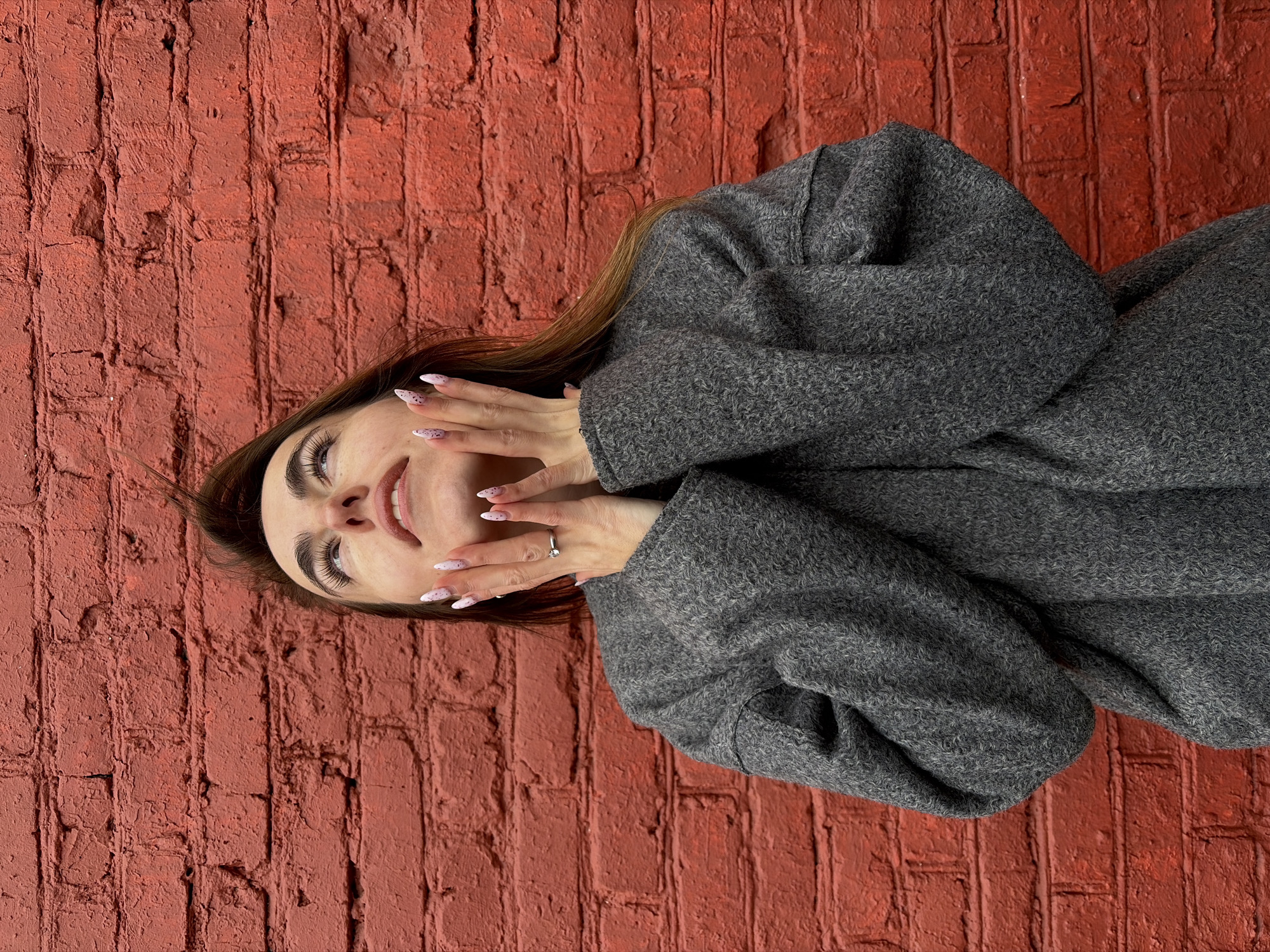 A woman with brown hair and manicured nails is lying on a red brick wall. She is wearing a gray coat and has her hands near her face. A ring is visible on her finger. The location is potentially Izhevsk, Russia.