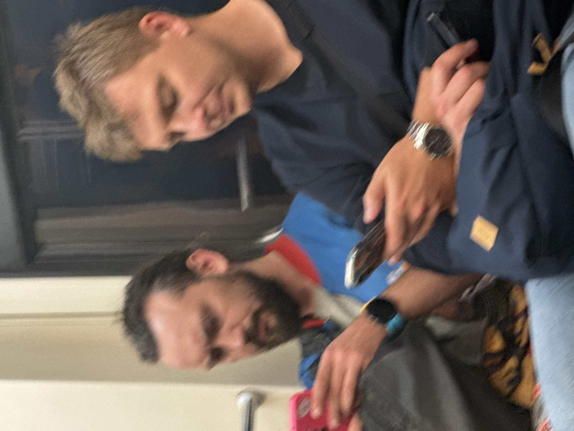 Two men are seated side-by-side, both looking down at their mobile phones. The man on the left has dark hair pulled back, a beard, and is wearing a blue and red jacket over a grey shirt. He is holding a pink phone in his left hand and a silver phone in his right hand. The man on the right has lighter brown hair and is wearing a dark blue long-sleeved shirt. He is holding a phone in his right hand and has a watch on his left wrist. They appear to be on a public transport vehicle, possibly a train, given the blurred background and seating arrangement. The overall lighting suggests an indoor setting. The location is not specified in the image.