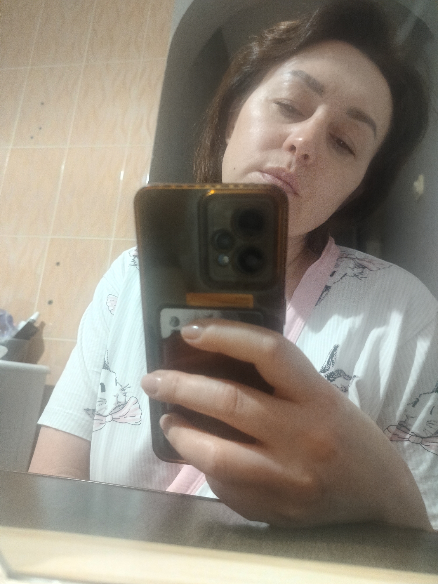A woman is taking a selfie in what appears to be a bathroom. She is wearing a white robe with pink trim and a cat print. The phone in her hand has a dark case and multiple camera lenses. The bathroom has light beige tiled walls and a mirror. The location is Chaikovsky, Russia.