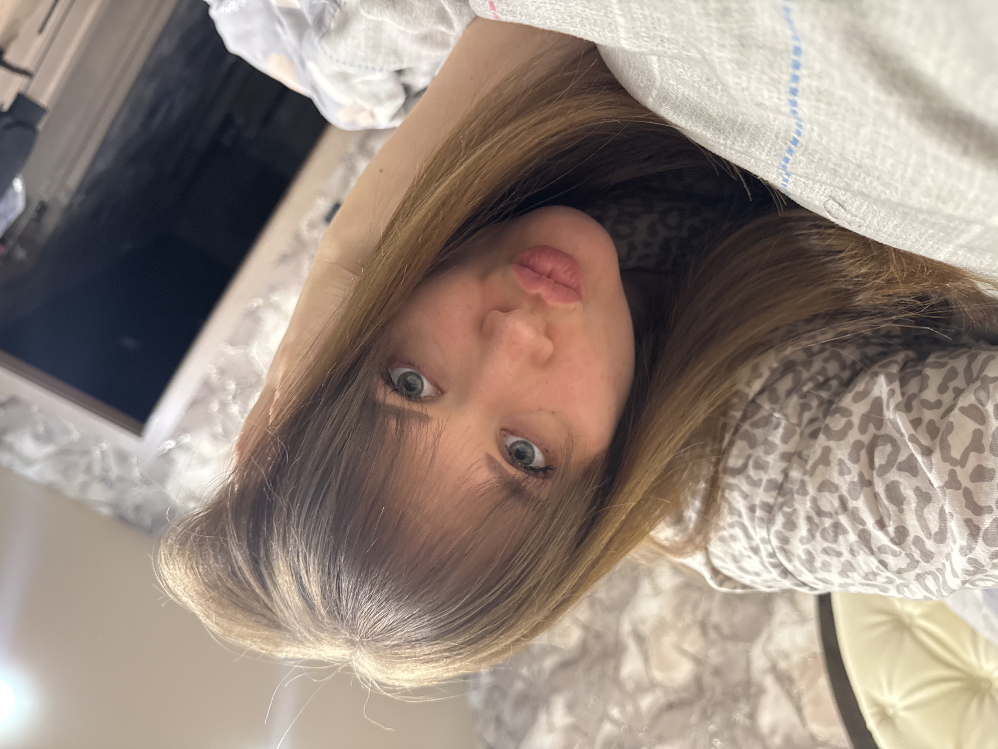 An indoor selfie features a young woman with light brown hair and light-colored, possibly blue or green, eyes. The image is oriented with the subject appearing on her side, rotated counter-clockwise. She gazes directly at the camera with wide, alert eyes and slightly pursed lips, conveying a playful or pouty expression. Her hair, with lighter highlights, frames her face, and some bangs fall over her forehead.
She is partly covered by a light-colored blanket or sheet featuring faint blue stripes, visible near the top of the frame. Her shoulders and parts of her arm are clad in a light gray top with a subtle leopard print pattern. The woman appears to be in a relaxed, possibly lying down, position.
The background is dimly lit and blurred, suggesting an indoor setting like a bedroom. It includes dark, reflective surfaces that could be furniture or a window, along with indistinct light sources. In the lower right corner, a light-colored, possibly upholstered object, resembling a headboard or furniture, is visible. There is no visible text in the image. The overall atmosphere is intimate and casual. The provided context indicates the location is Chaikovsky, Russia.