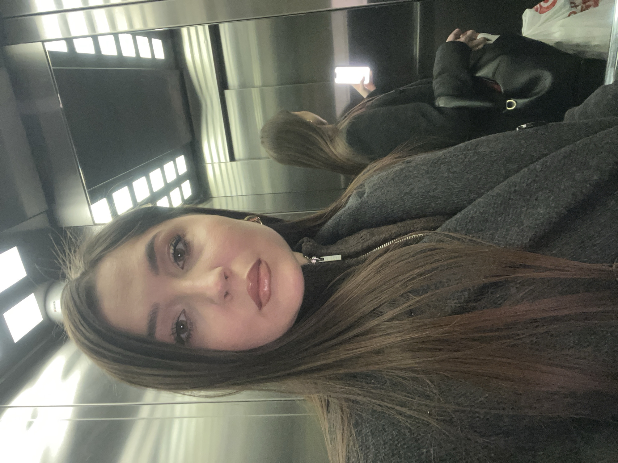 A selfie captures a young woman inside an elevator with highly reflective, metallic walls. The woman, positioned prominently in the foreground, has long, straight, dark brown hair, dark eyebrows, and a calm, neutral expression. She is wearing a dark grey or charcoal-colored knitted jacket with a prominent metallic zipper; the zipper pull bears faint text that appears to begin with "VERSA". Her lips are adorned with a light-colored, glossy lipstick.
The elevator's interior features artificial lighting from horizontal rectangular fixtures, which are clearly reflected in the polished wall surfaces. In the background reflections, the woman is seen from behind, holding a brightly glowing smartphone, indicating she is actively taking the selfie. Adjacent to her reflection, a black bag or purse with a visible gold D-ring is present. A white plastic bag with a distinctive red bullseye logo, strongly resembling the Target brand, is also partially visible in the reflection.
The image depicts an indoor scene with artificial light, preventing any determination of the time of day or weather. The activity is a woman taking a mirror selfie. The provided context indicates the location is Khimki, Russia.