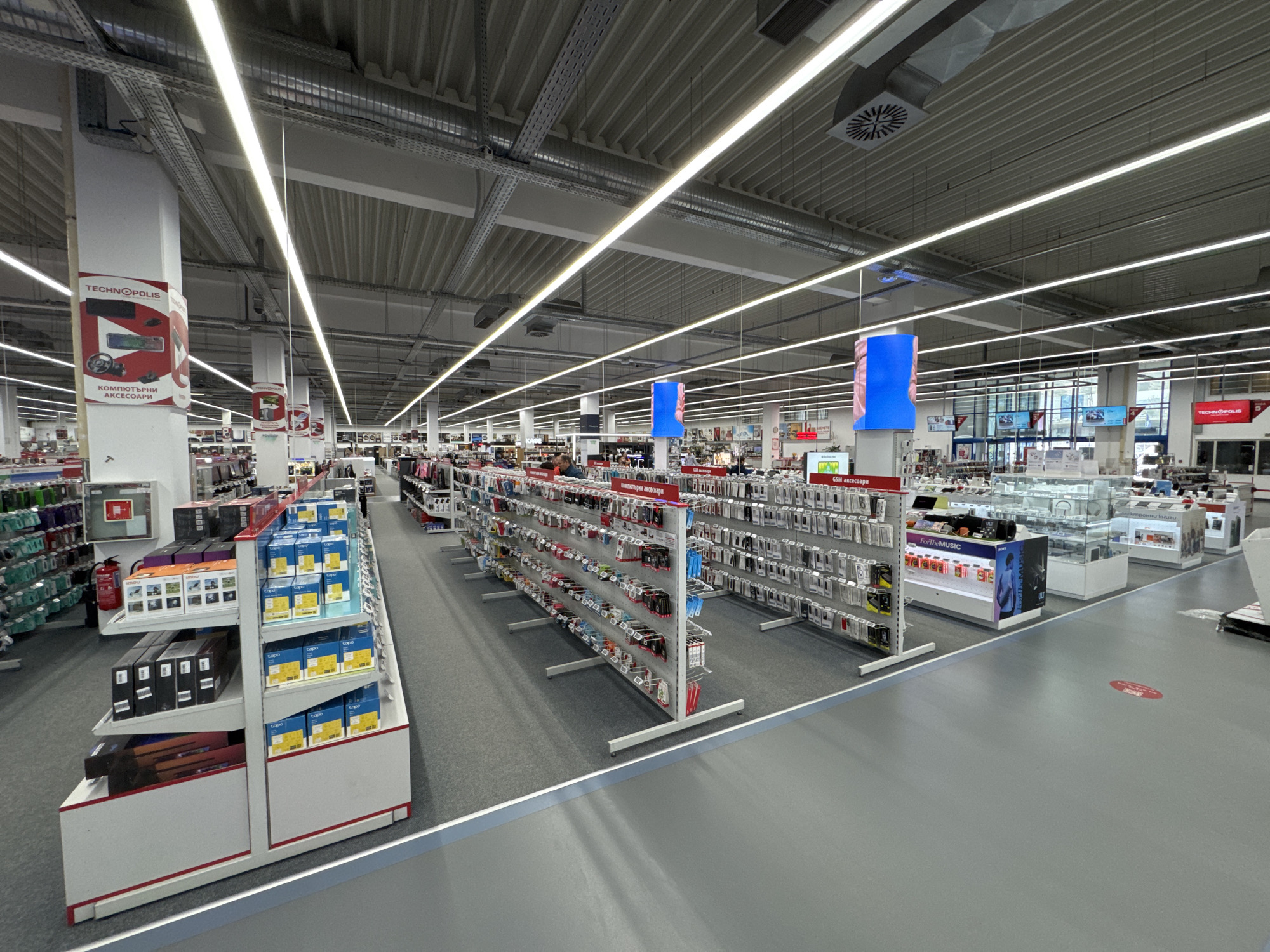 The image shows the interior of a TECHNOPOLIS store in Sofia, Bulgaria. The store is well-lit with overhead fluorescent lights and has a high, corrugated metal ceiling with visible ventilation systems. The floor is a combination of grey carpet and grey linoleum. Aisle shelves are stocked with various consumer electronics and accessories, including computer accessories and GSM accessories. The shelves are organized and labeled. There are several white display units and signage with brand logos. Aisle markers with blue tops indicate the categories of items for sale. Customers are visible in the background browsing the merchandise. A red fire extinguisher is visible mounted to a pillar. A sign on a pillar shows the logo of TECHNOPOLIS along with the words "КОМПЮТЪРНИ АКСЕСОАРИ". A red round sticker is placed on the floor.