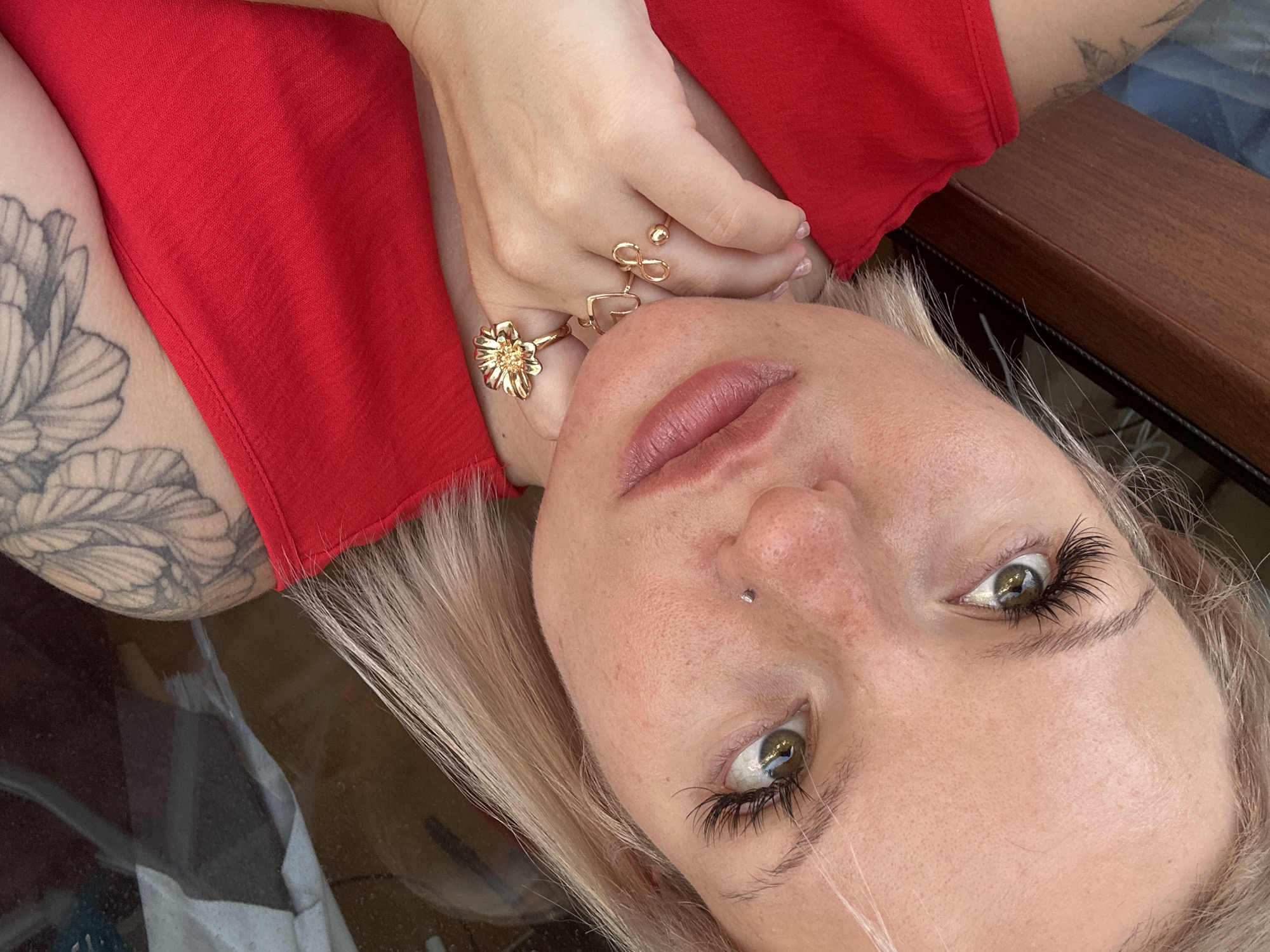 The image shows a close-up, upside-down, low-angle view of a person's face, specifically their right eye, nose, and mouth. They have light-colored hair and large, dramatic eyelashes. A small silver stud is visible on their nose. The person is wearing several rings on their fingers, including one shaped like a flower and another resembling an infinity symbol. They are wearing a bright red garment, possibly a top, and have a tattoo on their left arm, partially visible. The background appears to be a wooden surface and possibly glass, suggesting an indoor setting or a place with furniture. The lighting suggests it could be daytime, with a soft, natural quality. The overall mood appears relaxed, with the person looking directly into the camera. There is no visible text in the image.