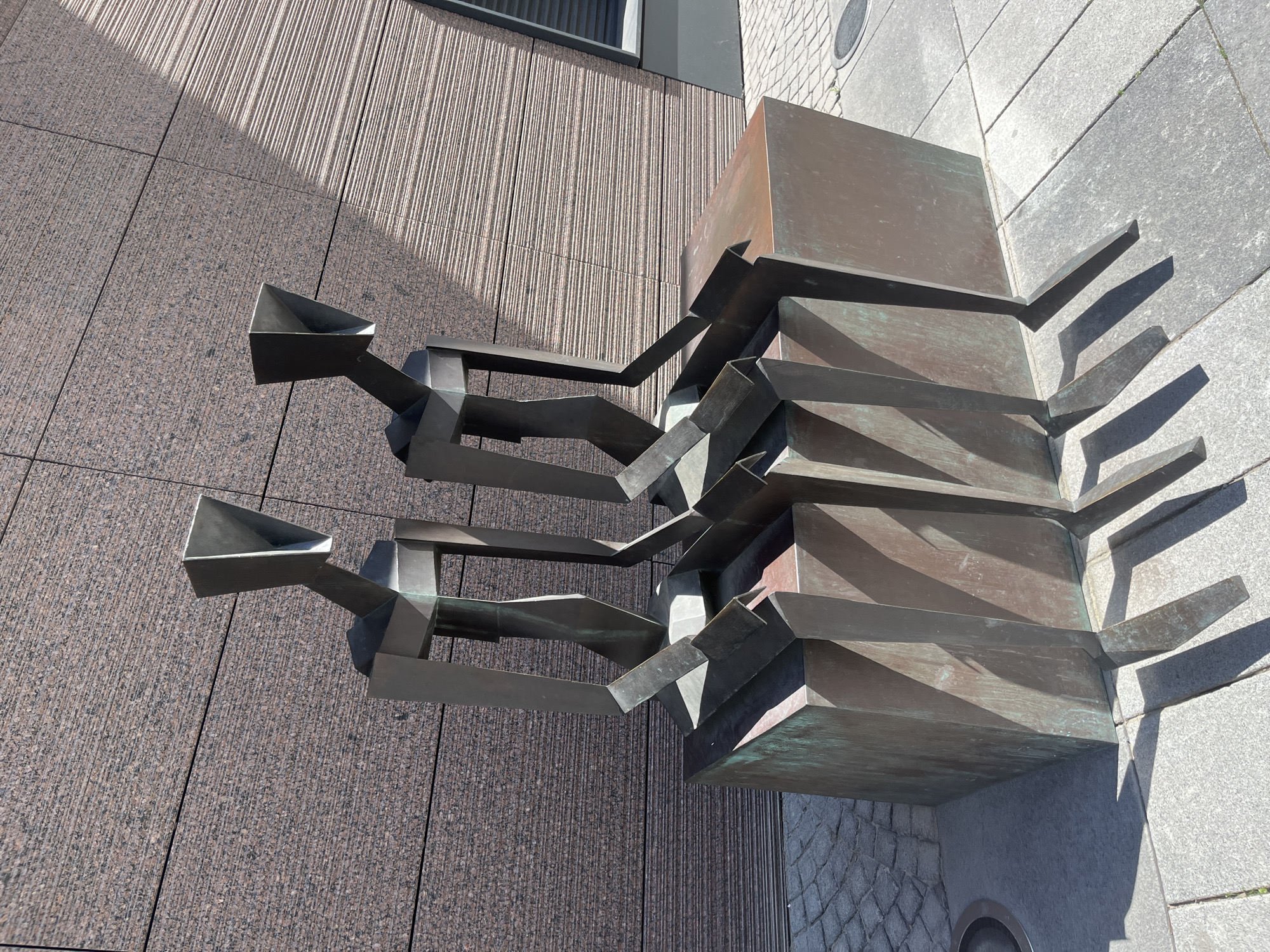 This image shows a modern metal sculpture with a geometric design, likely made of bronze, situated outdoors next to a building with a granite facade. The sculpture has a series of stacked rectangular elements, with protruding, angled arms. The overall impression is abstract and industrial. Two of the angled arms have funnel-like openings.
The sculpture is positioned on a paved area with large stone tiles, and a darker textured pavement is visible to the right, possibly cobblestone. The building's wall is made of large, dark granite tiles with vertical textured lines, creating a rhythmic pattern. A shadow of the sculpture falls across the pavement, indicating it is daytime. The bright sunlight suggests a clear day.
No people are visible in the image. The scene appears to be a public space, likely in a city, given the architecture and urban paving. The materials and design suggest a contemporary setting. There is no visible text in the image. The lighting and shadows suggest the photograph was taken around midday. The location context provided is Dnipro, Ukraine.