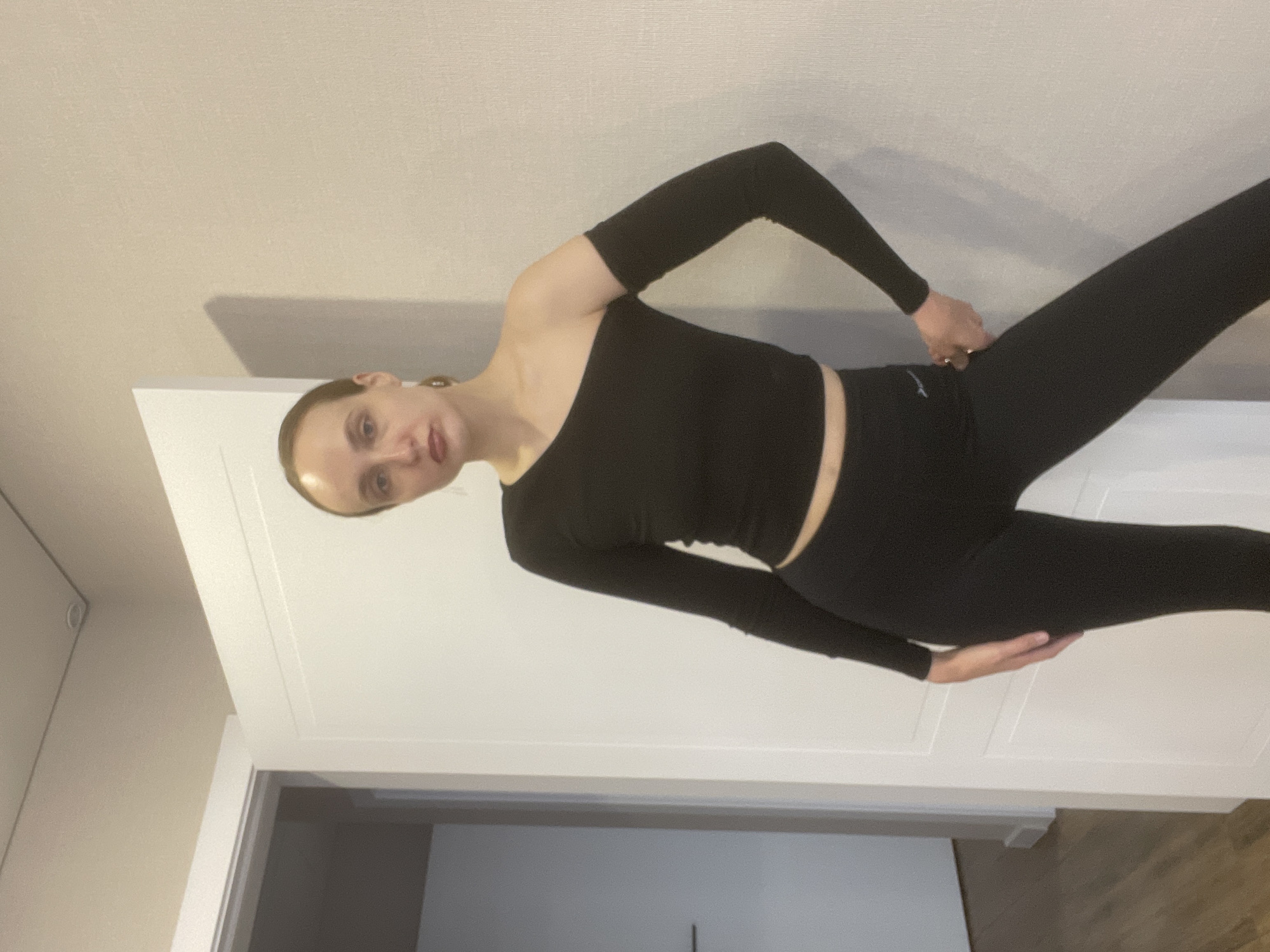 A young adult female is captured in a full-body indoor portrait, standing in what is likely a home environment in Подшивалово, Russia. The subject is wearing an all-black athletic or casual ensemble, featuring a long-sleeved, one-shoulder crop top and matching full-length leggings. Her hair is pulled back, and she gazes directly at the camera with a subtle, neutral smile.
She poses with her right arm bent, hand resting near her hip, and her left hand gently placed on her left thigh. A small, indiscernible white logo or text is visible on the upper left thigh of her leggings. The background primarily consists of a white paneled door or cabinet, filling a large portion of the frame behind her. Above this, a plain, light beige wall is visible, with soft shadows suggesting indoor artificial lighting. The scene conveys a relaxed, casual moment.